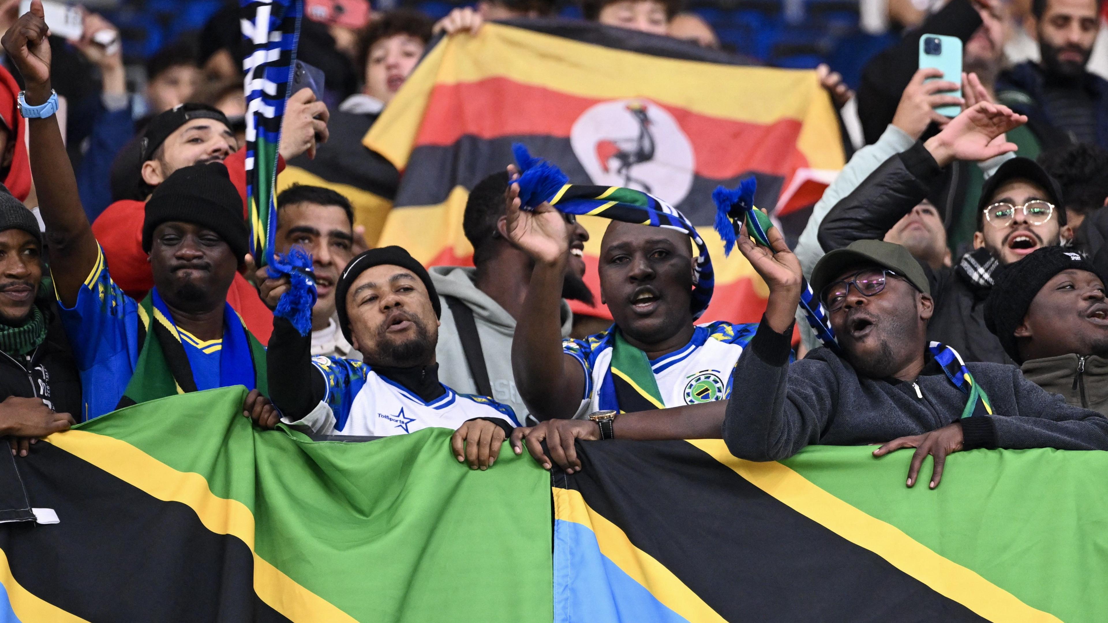 A group of fans with flags display their allegiances in the stand at the Afcon 2025 match between Tanzania and Uganda. A front row of Tanzania fans, three wearing Tanzania jerseys, drape two Tanzanian flags over a barrier in front of them. More fans can be seen dancing and waving their hands behind them