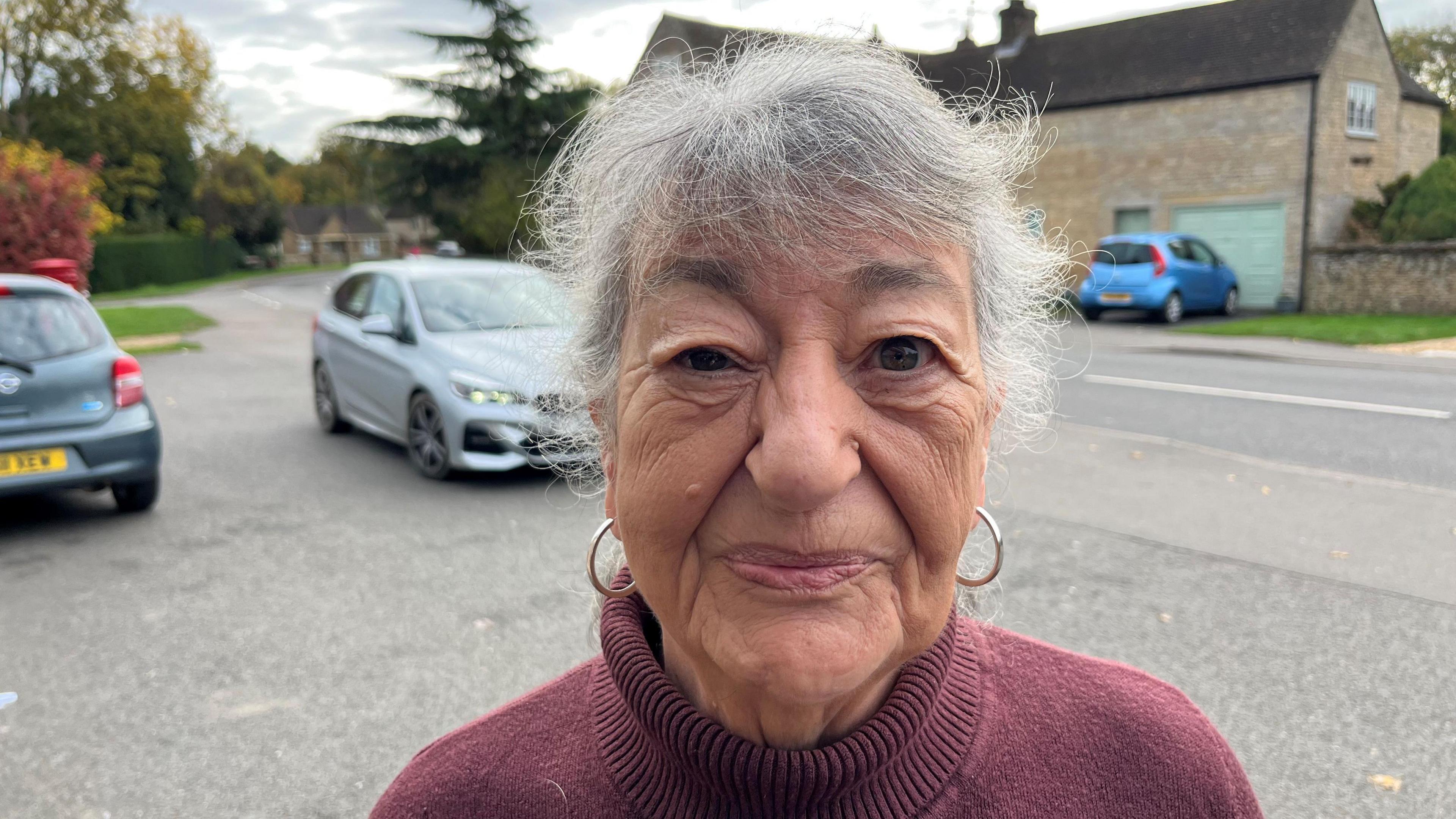 Marisa Clifton has short grey hair over her forehead and two large gold earrings she is wearing a turtle neck maroon woollen jumper. Behind her are some parked cars and a property visible too.