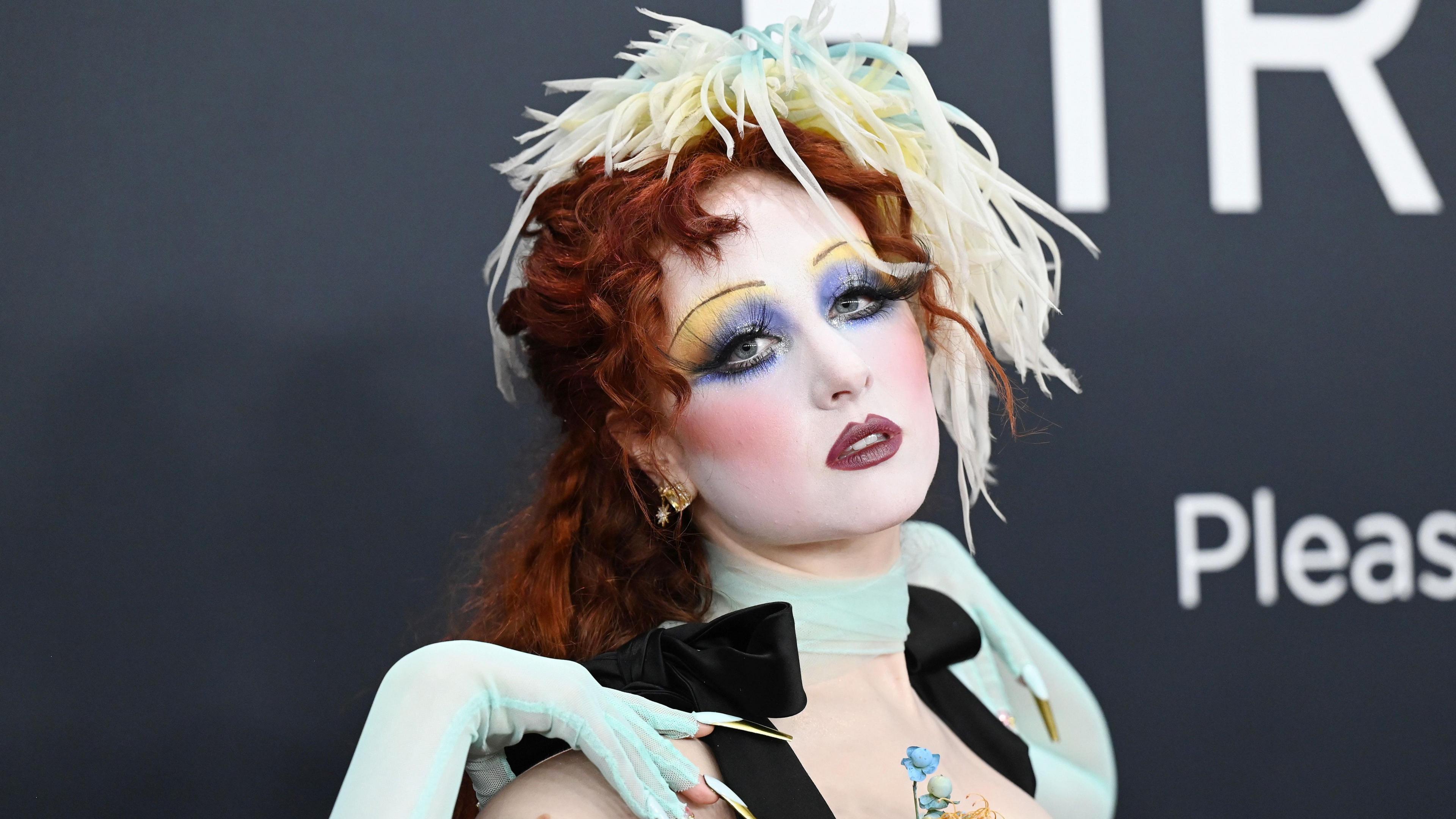 Chappell Roan on the red carpet for the Grammy Awards. She wears pale blue gloves which match a scarf tied tightly around her neck and a hair accessory which is also pale yellow. She wears elaborate make-up including pencil thin brows and yellow and purple eyeshadow. She poses with her head tilted back and her right hand rested on her shoulder. 