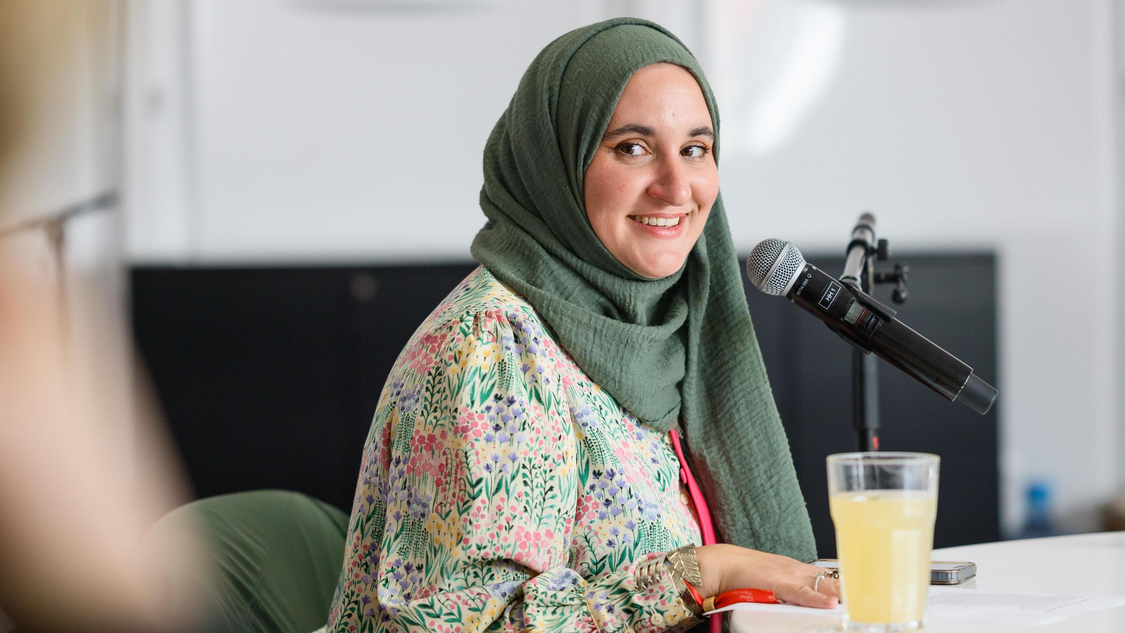 Hanan sat at a table with a microphone in a stand. She is turning to smile at the camera and is wearing a top with flowers on it and a sage green headscarf.