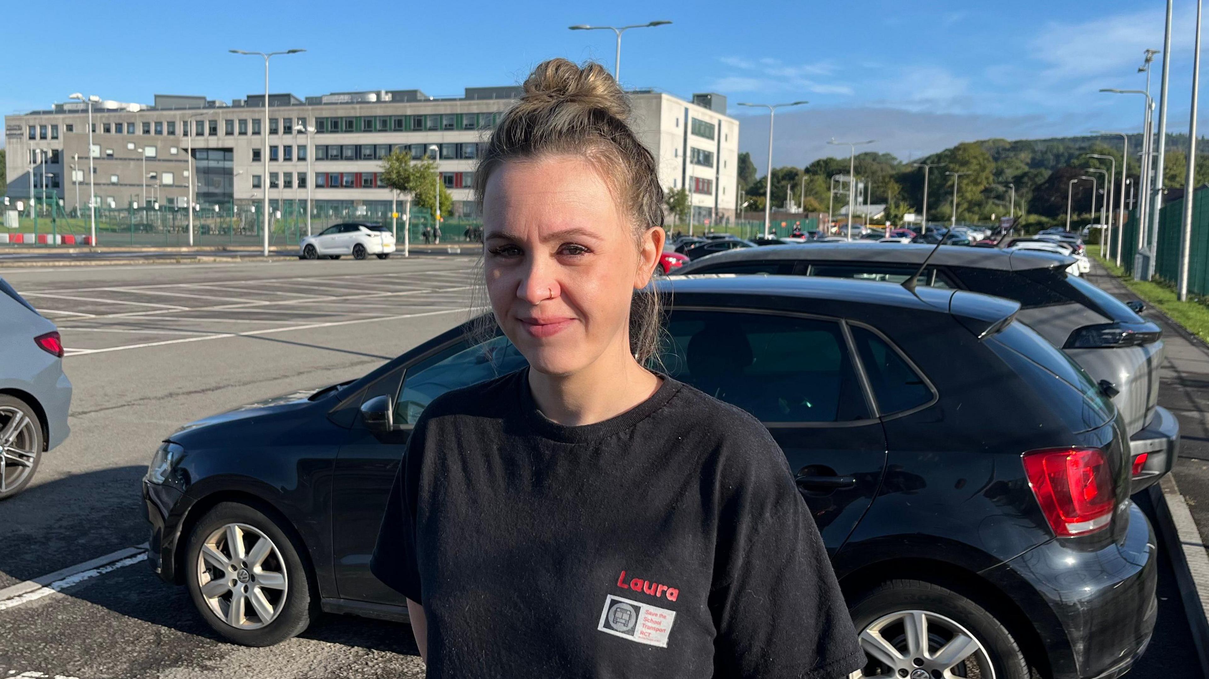 A woman with brown hair, wearing a black t-shirt, is seen standing in a car park