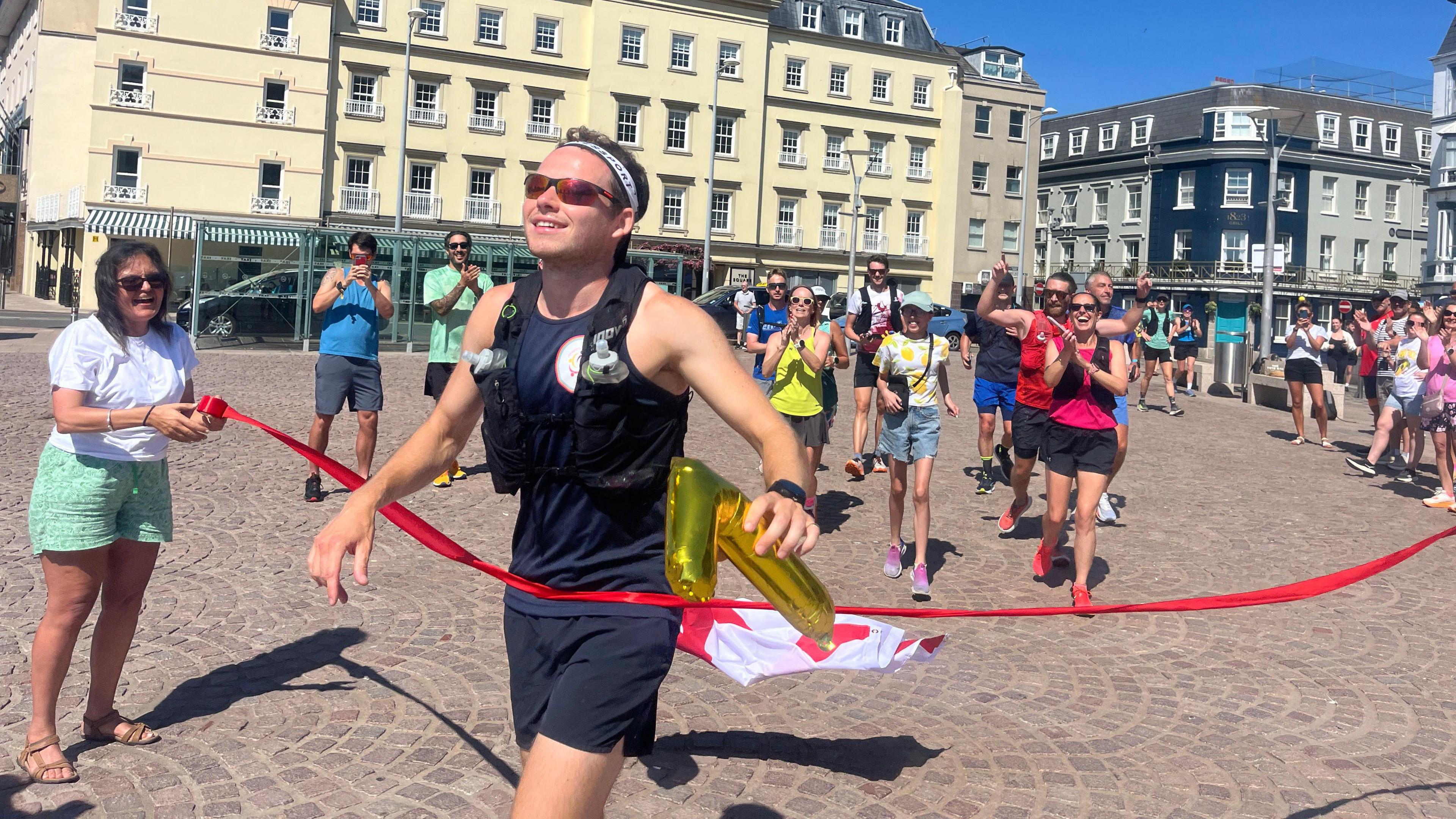 James crosses the finish line of a marathon in an urban square, triumphantly holding a golden inflatable number '7'. A red ribbon marks the finish, held by another person. Behind James, other participants cheer and clap, with tall buildings and a clear blue sky in the background