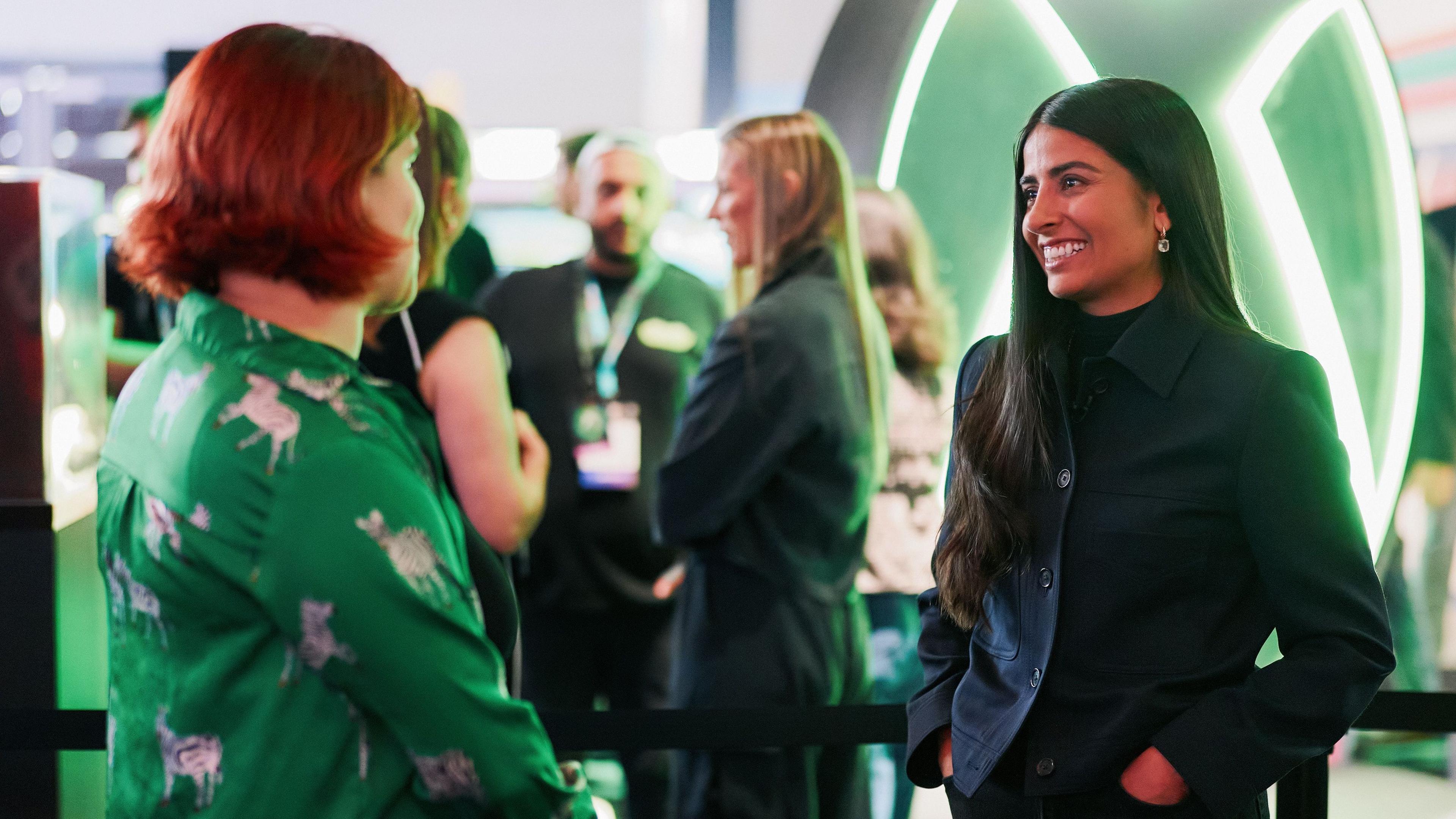 Two women stand in a convention hall with a big green Xbox logo behind them. On the left, a person with a green shirt and short red hair. On the right, a woman with long brown hair and a dark jacket.