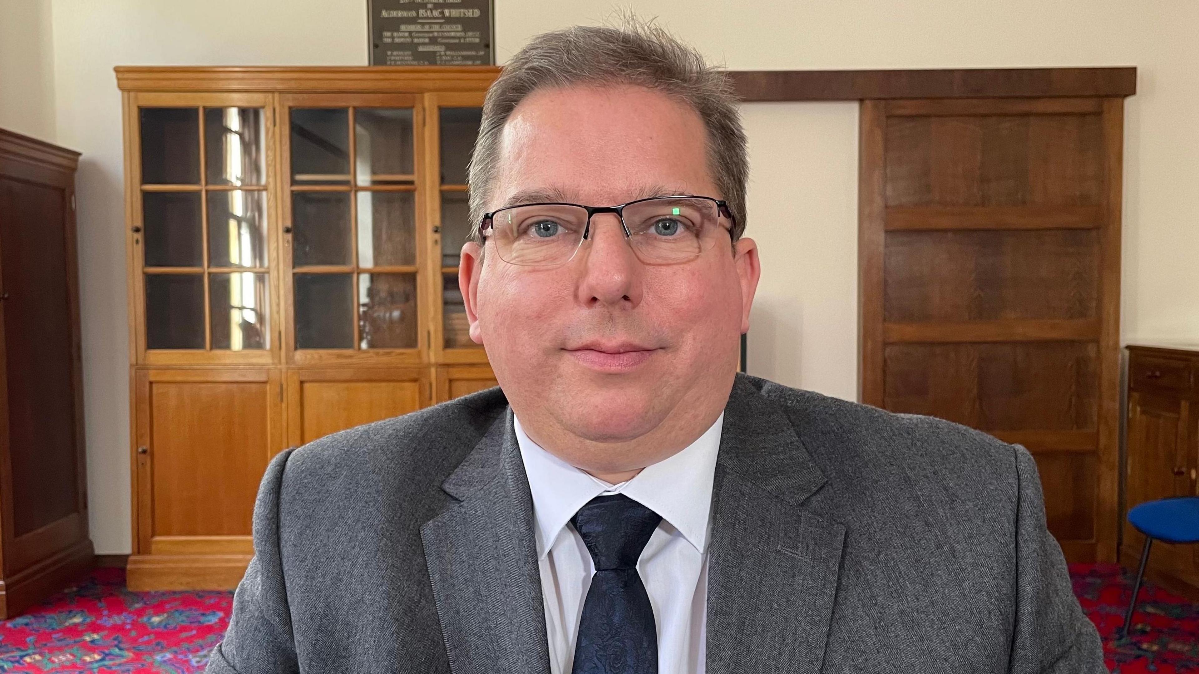 John Gregg, looking direct to camera. He is wearing a grey suit, white shirt and navy blue tie. He is clean shaven, with glasses, blue eyes and light brown hair. He is sitting in the middle of a large room and behind him are an empty, glass-fronted bookcase, and a wooden door panel. Light is coming from the left of the image, but the windows are out of the frame, though one can be seen reflected in the glass of the bookcase. A heavily-patterned red and blue carpet can just be seen in the background.