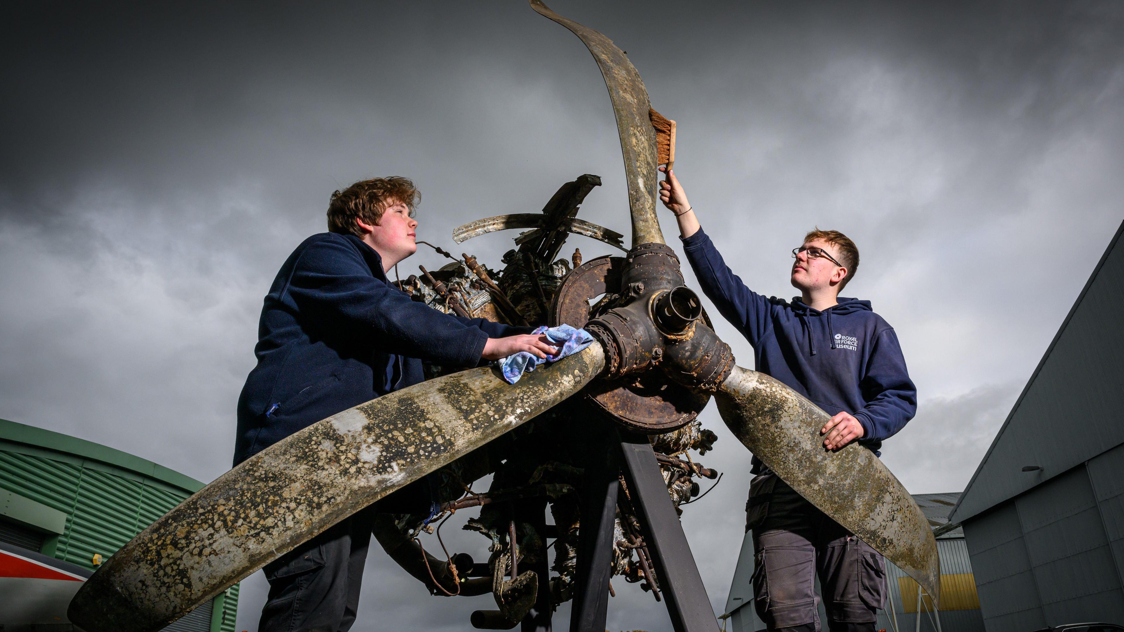 Two young men in navy RAF Museum hoodies are standing next to a large propeller from a war plane. It is metal and various shades of rust, brown and green. In the background, the sky is grey and cloudy