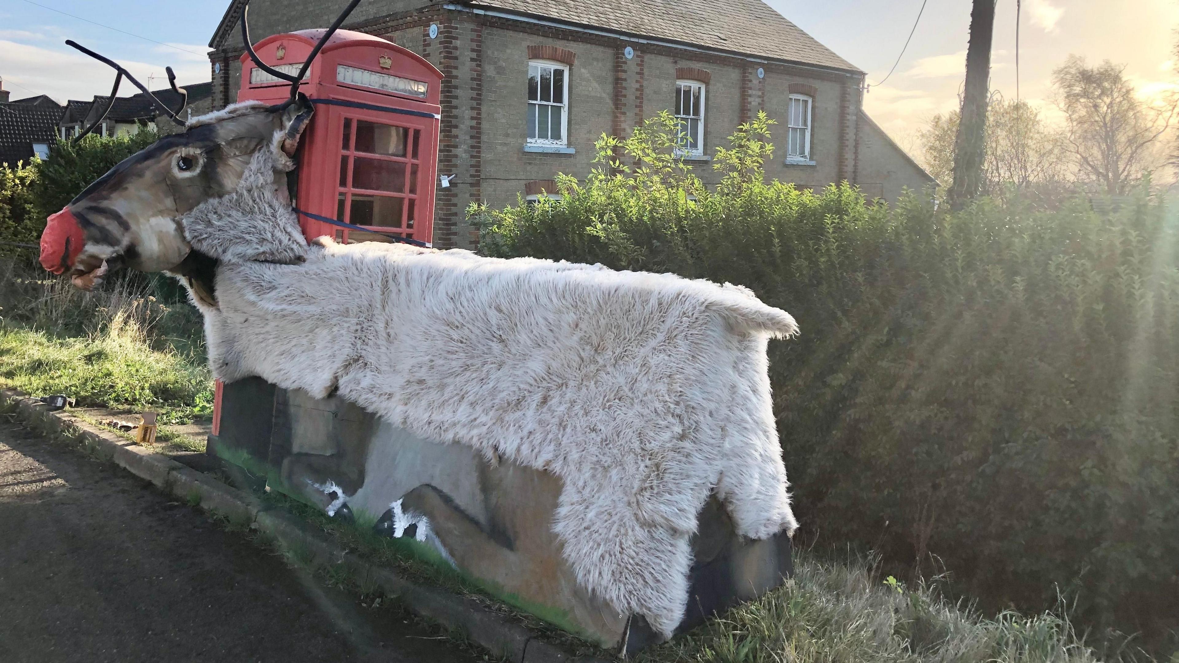 A sculpture of a reindeer attached to a telephone box. It has a large head made from fabric and its wooden body is covered in fake fur
