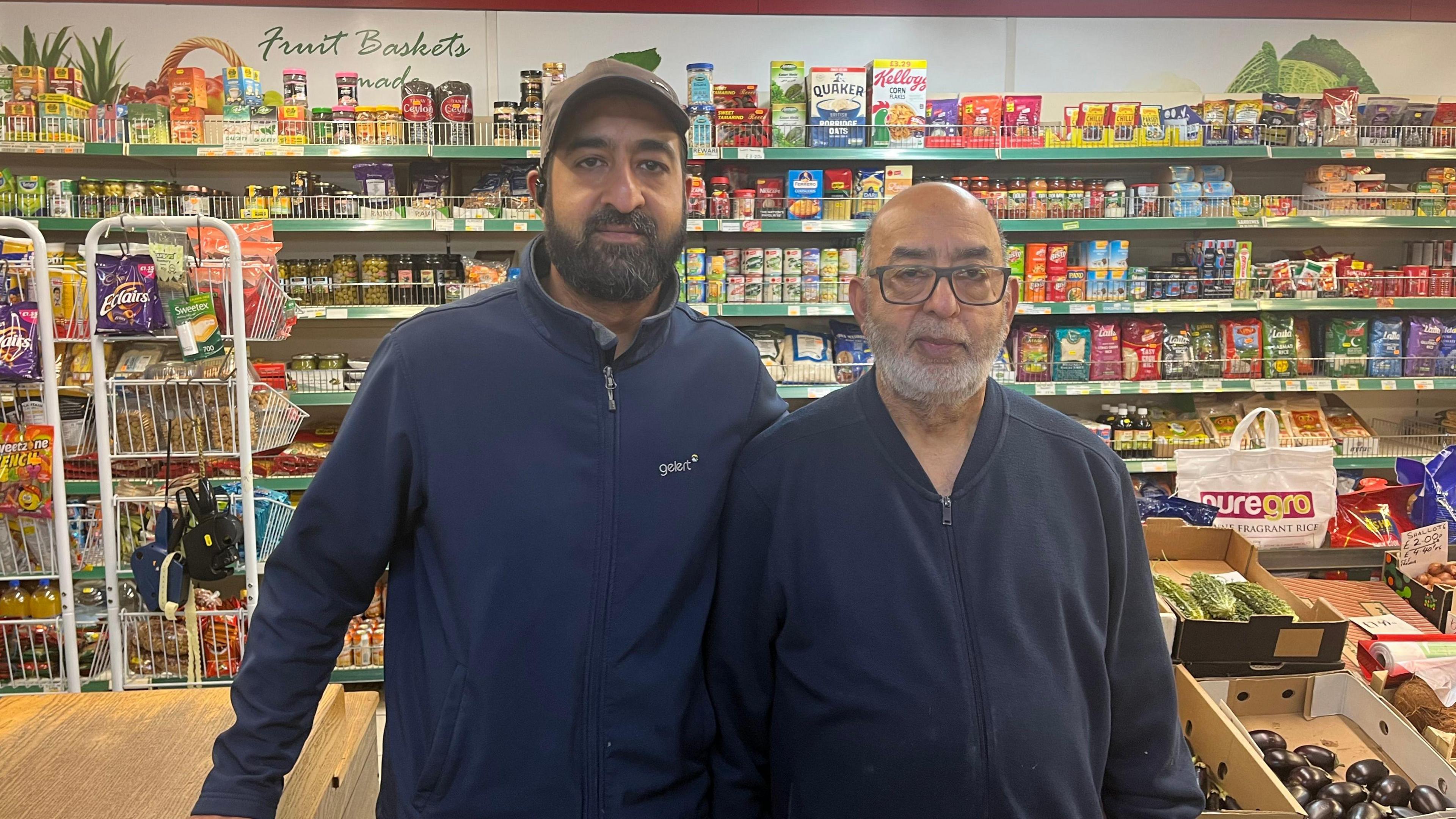 Two men, one on the right in his 60s, the one on the left in his 40s, standing in front of a shop