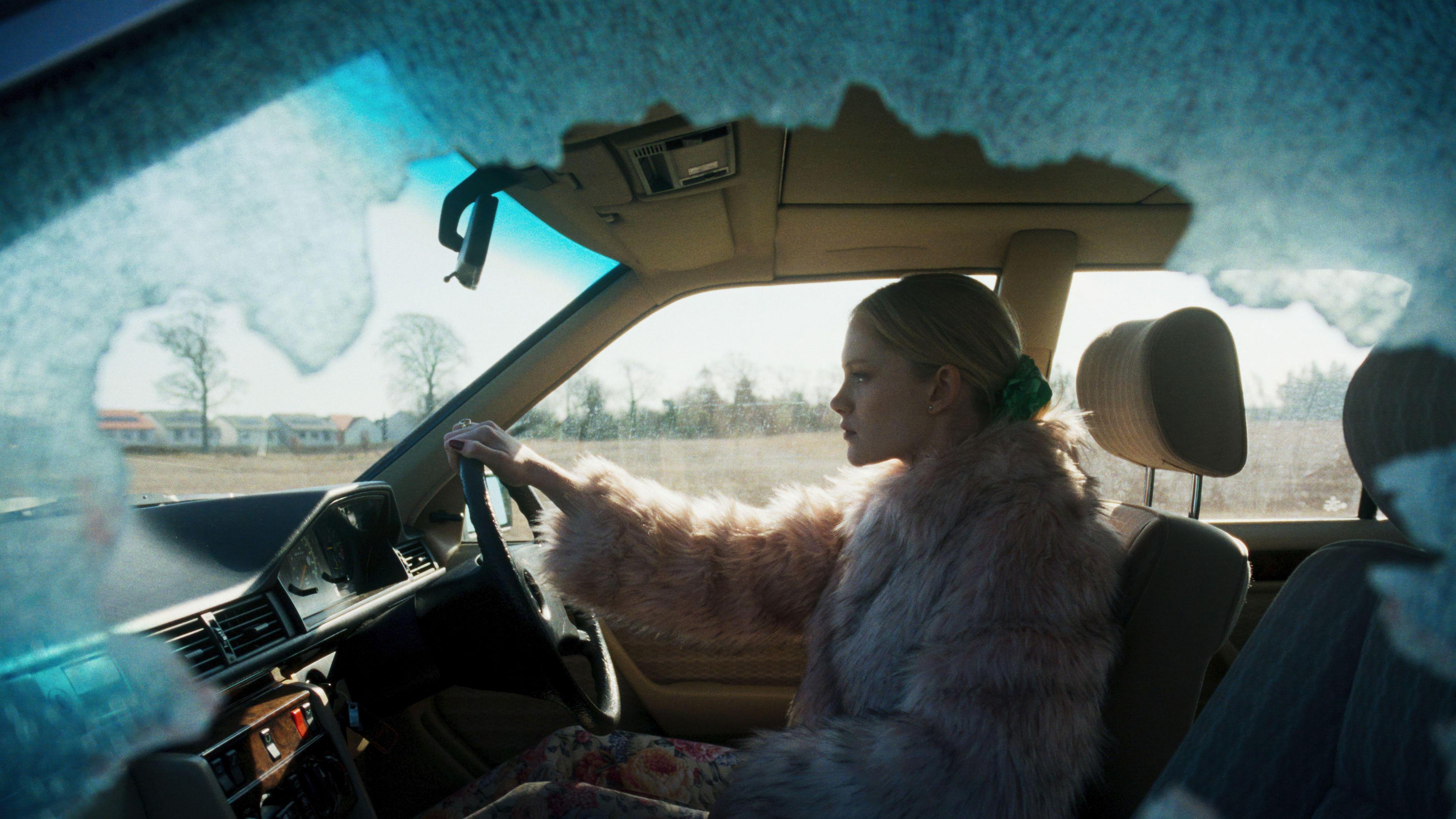 A woman wearing a pink furry coat drives a car with a smashed passenger side window