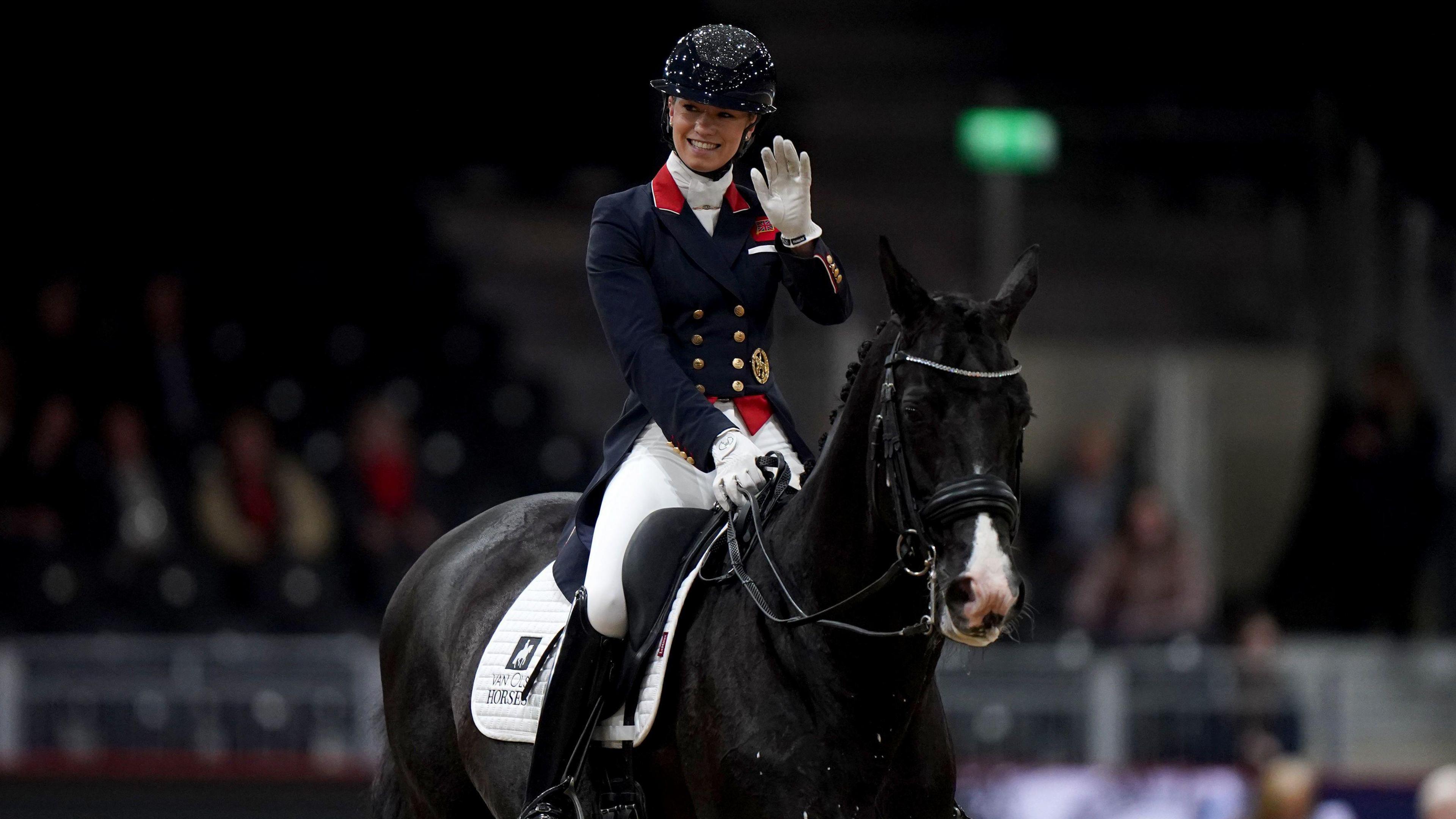 Lottie Fry, wearing blue and white riding clothes and a black helmet, smiles and waves as she rides a black horse at the  FEI Dressage World Cup in London in 2023.