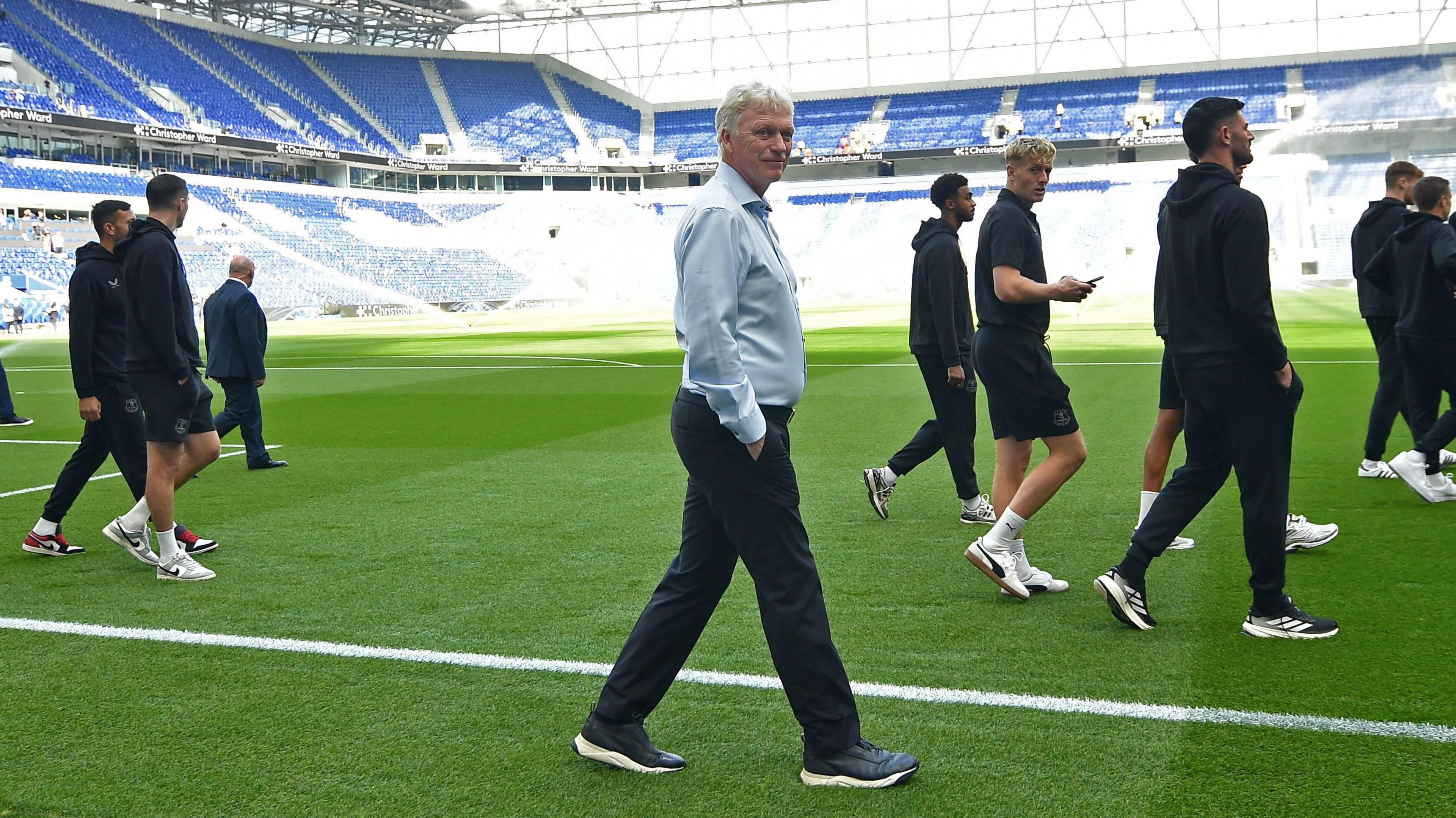 Side view as David Moyes, smiling in the centre and turning to camera, and about eight Everton players in front and behind him - who are in dark training gear - stroll around the green pitch before the game. Empty blue seats can be seen in the stands behind.