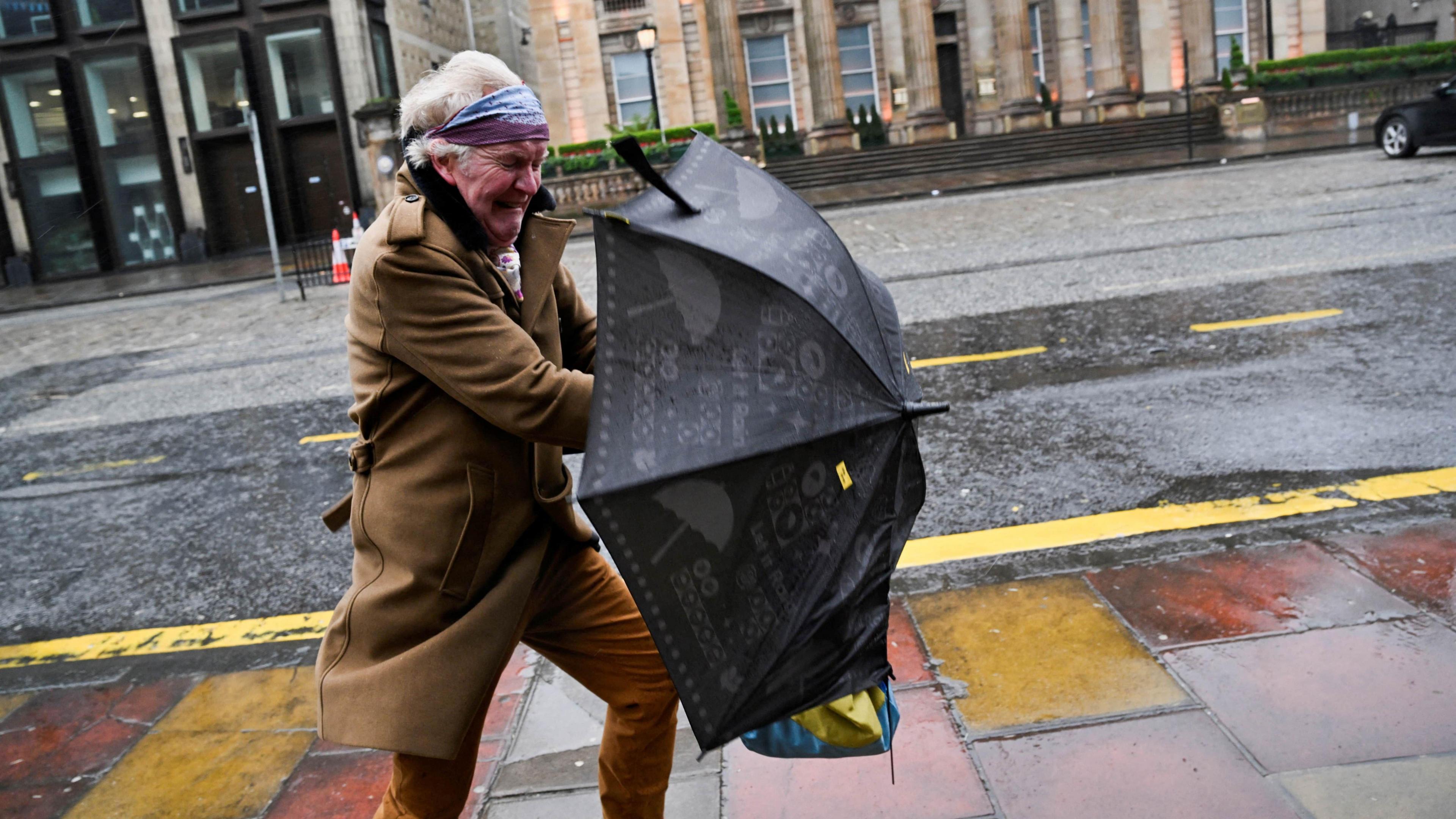 A woman struggles with an umbrella on George Street in Edinburgh. She is wearing a long brown coat with brown trousers and a multi-coloured head band. She is holding a black umbrella and grimacing against the wind.