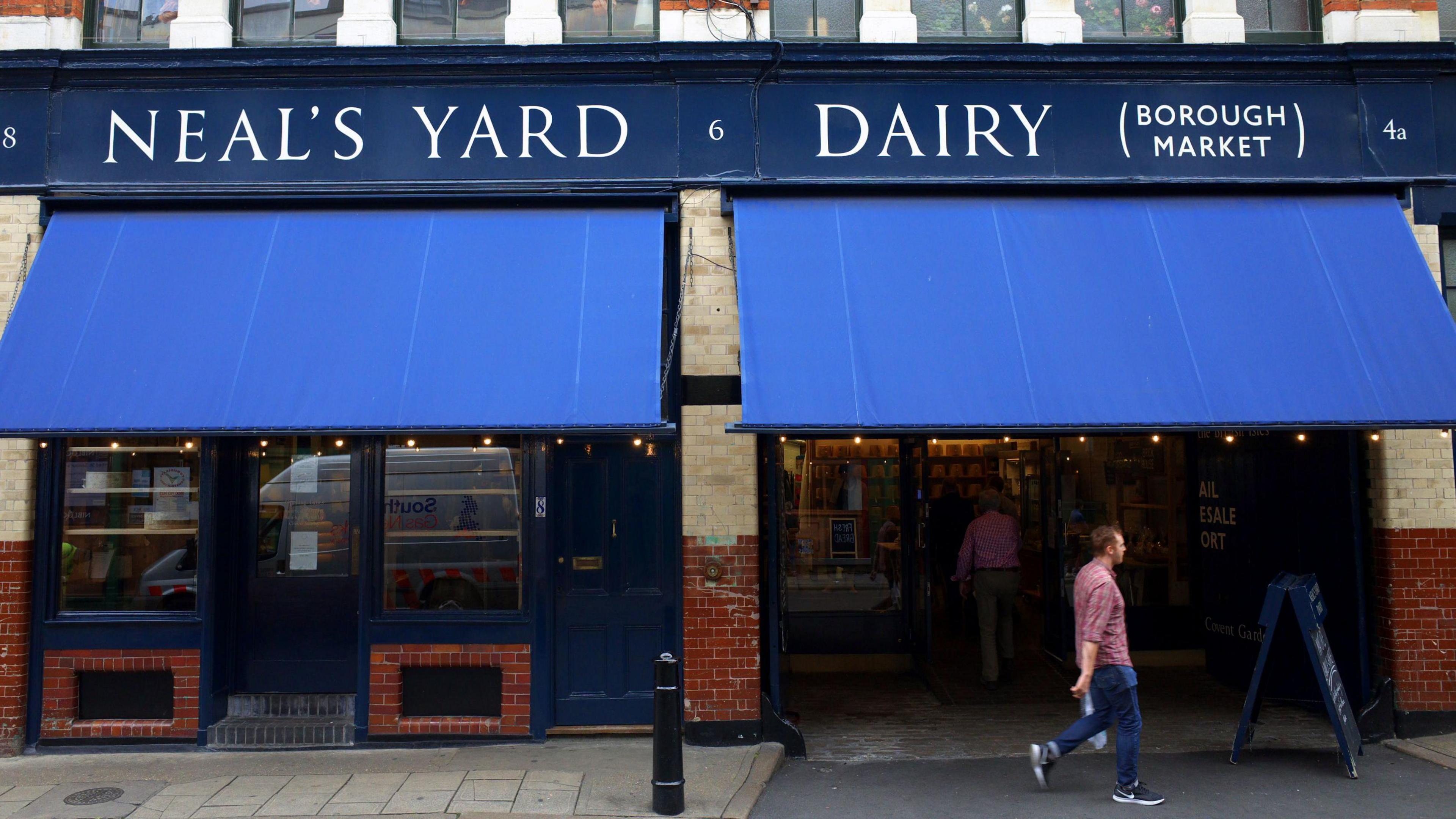 The outside of the store in Borough Market, has Neal's Yard Dairy written in white and blue sign with empty shelves in the front window and a man wearing a red chequed shirt and blue jeans walking past the right hand side of the store