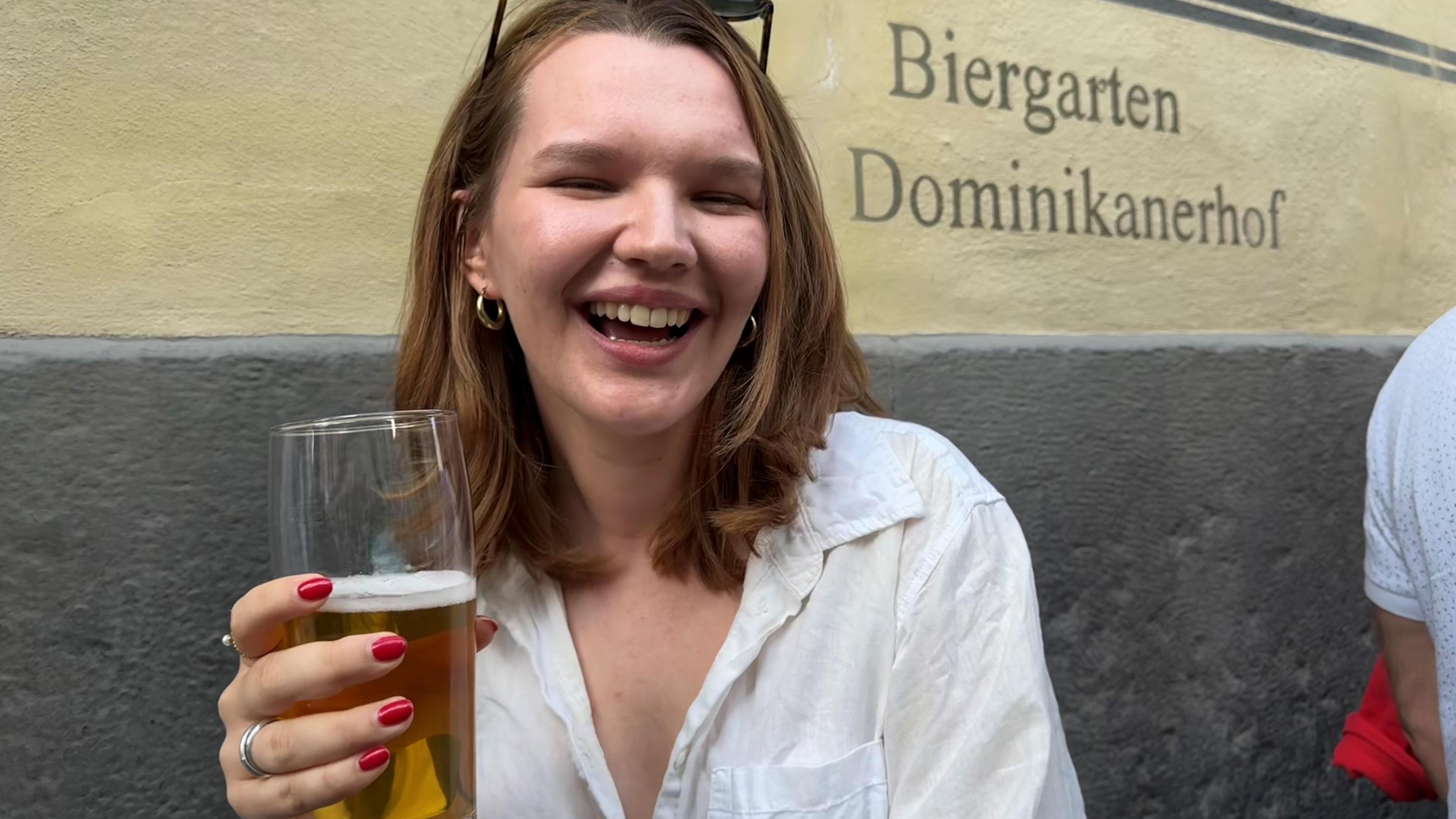 A young lady smiles to the camera holding a half-drunk beer. She has a long auburn bob and red painted nails.