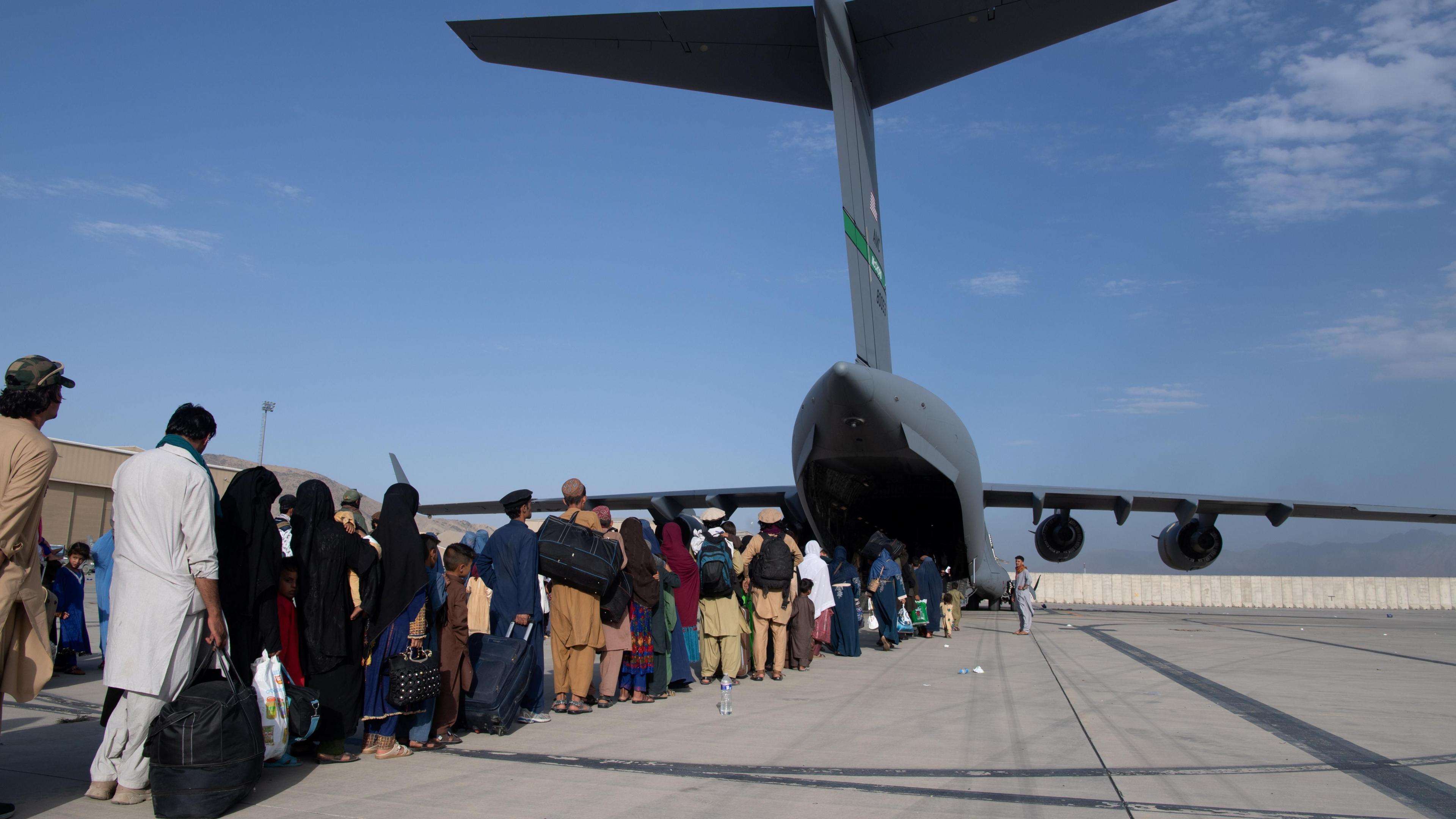A line of Afghan evacuees line up behind a military aircraft, holding large bags, suitcases, and babies. The women are covered with a headscarf, and wearing black, while the men wear colour. At the opening at the back of the plane, a member of military personnel counts people onto the plane.