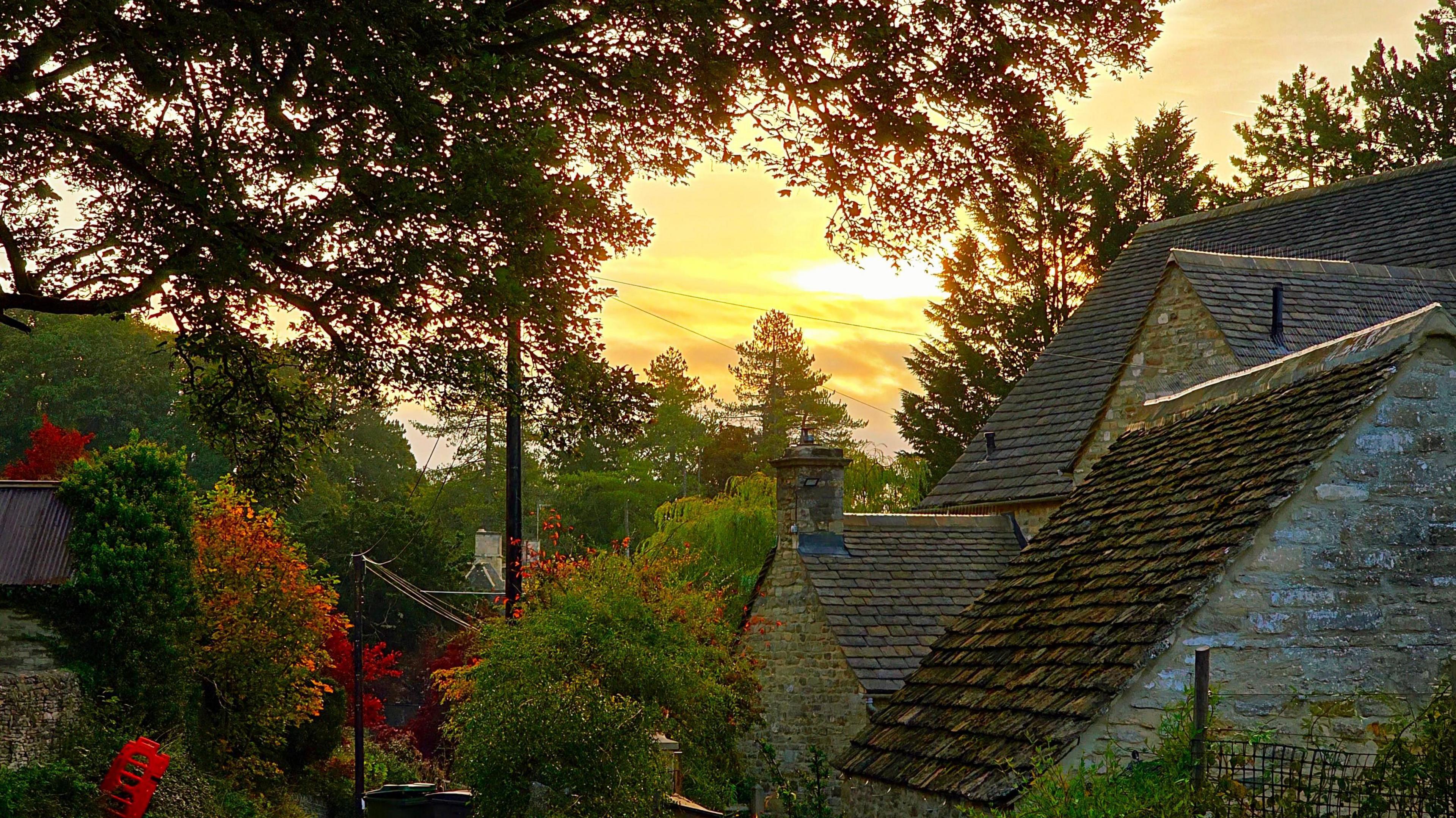 A pale, pink-hued watercolour sunrise is framed by a Cotswold stone honey coloured cottage and trees, one of which is bursting with red leaves.