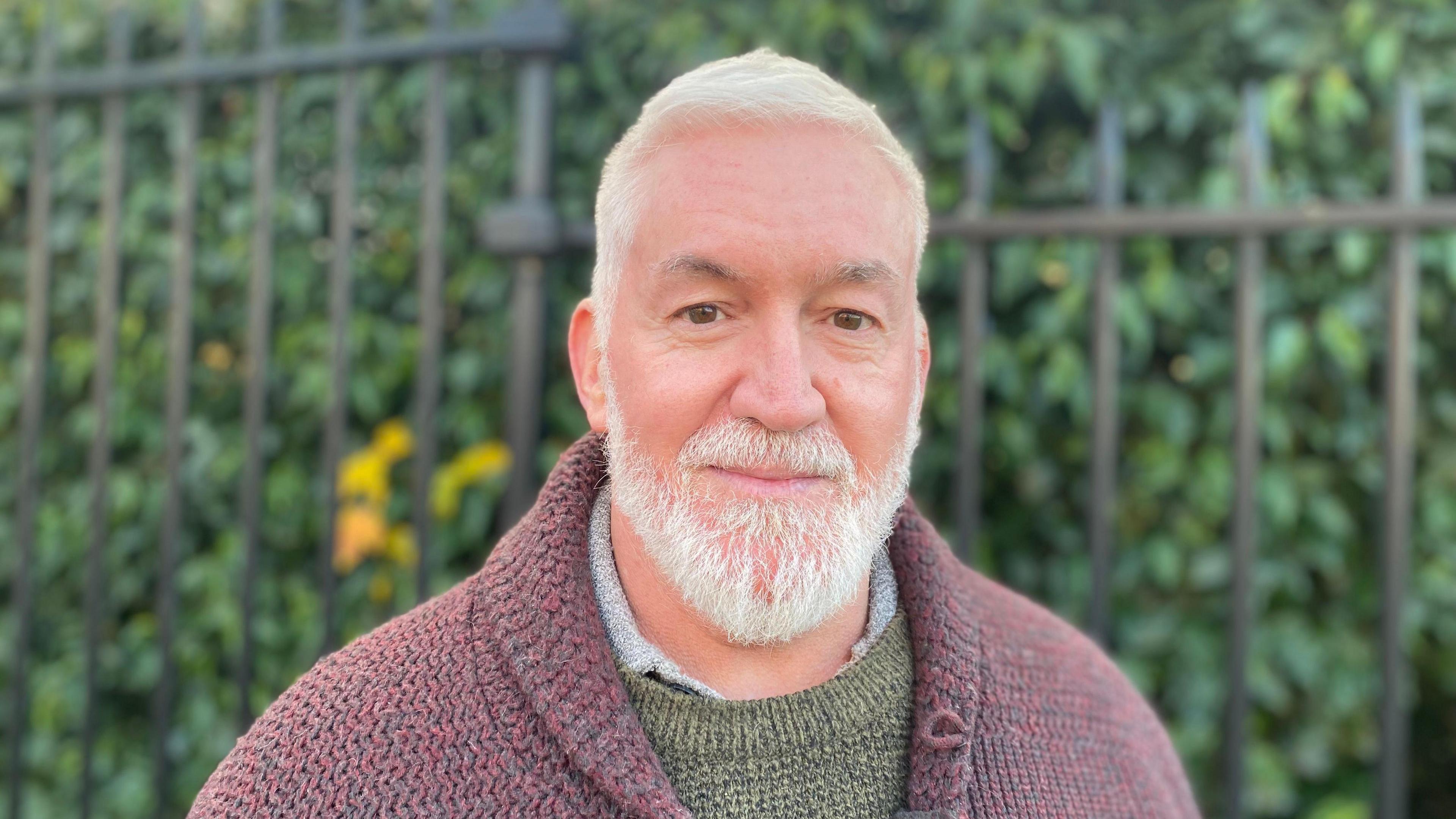 A man wearing a green knitted jumper under a burgundy knitted jacket. He's standing in front of green shrubs.