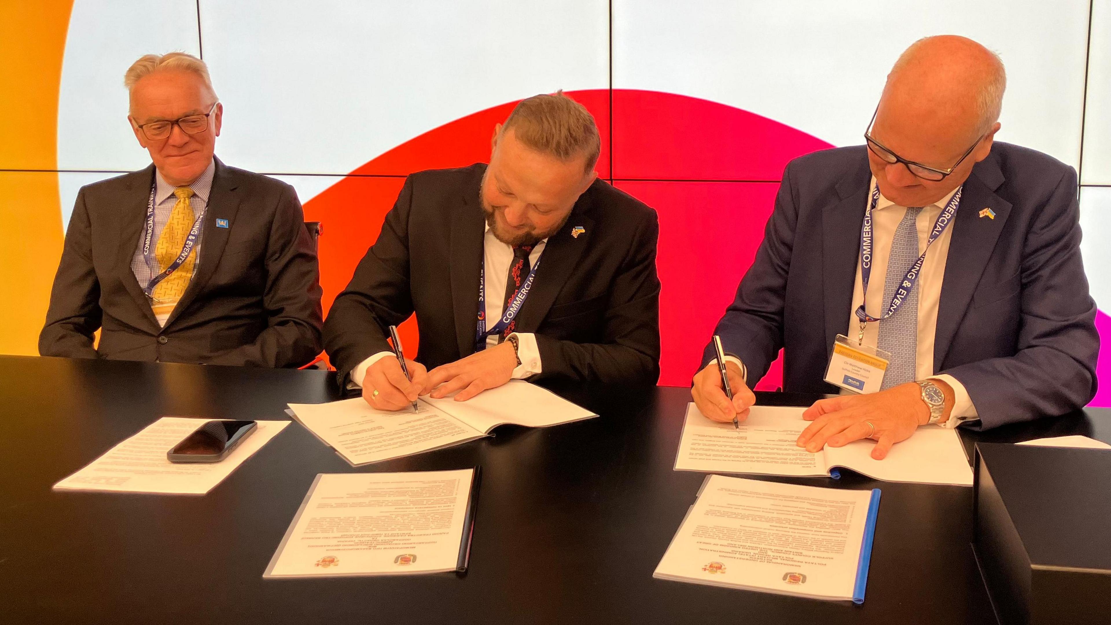 Three men sit at a black table. They are were wearing suit jackets and white shirts. Two of them are signing white pieces of paper.