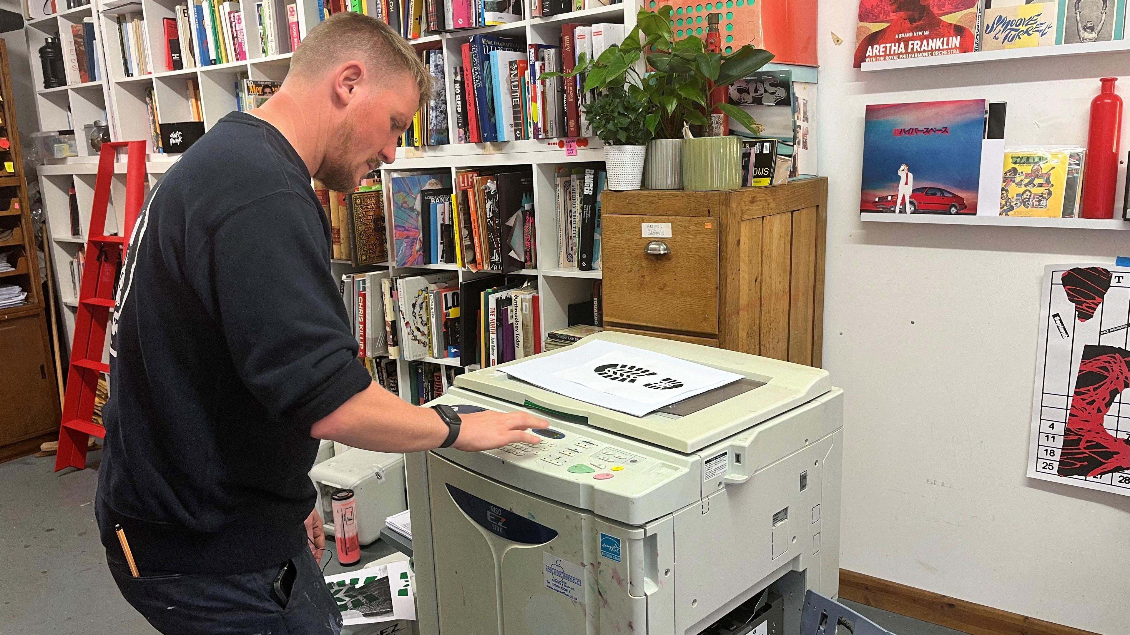 Jimmy Turrell loading a Risograph printer with images. It resembles a big printer and photocopier/scanner. There are plants on a wooden filing cabinet behind it.