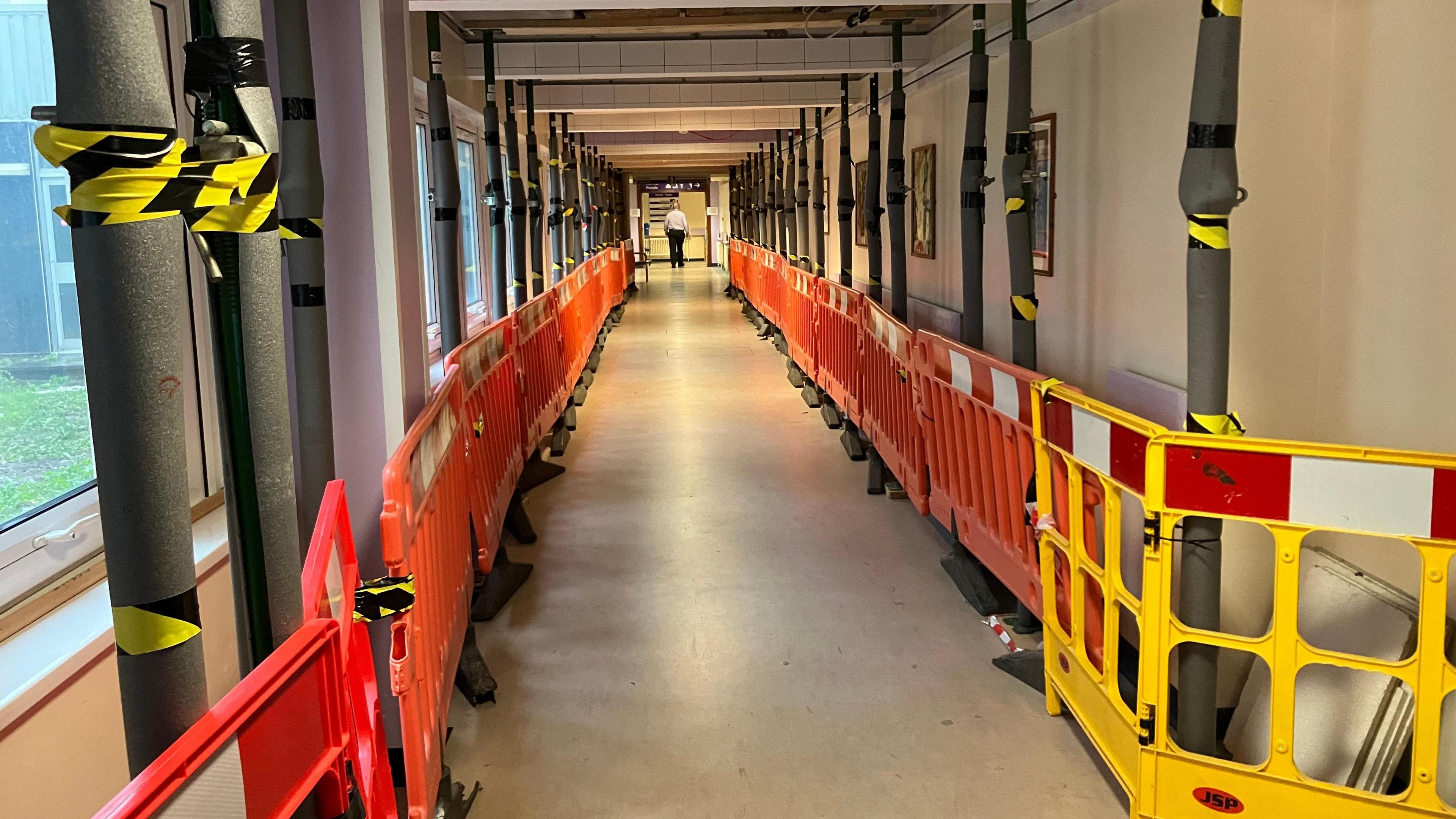 A hospital corridor is seen with yellow and orange barriers on either side, running the length of the corridor. Black and yellow tape is wrapped around pillars.