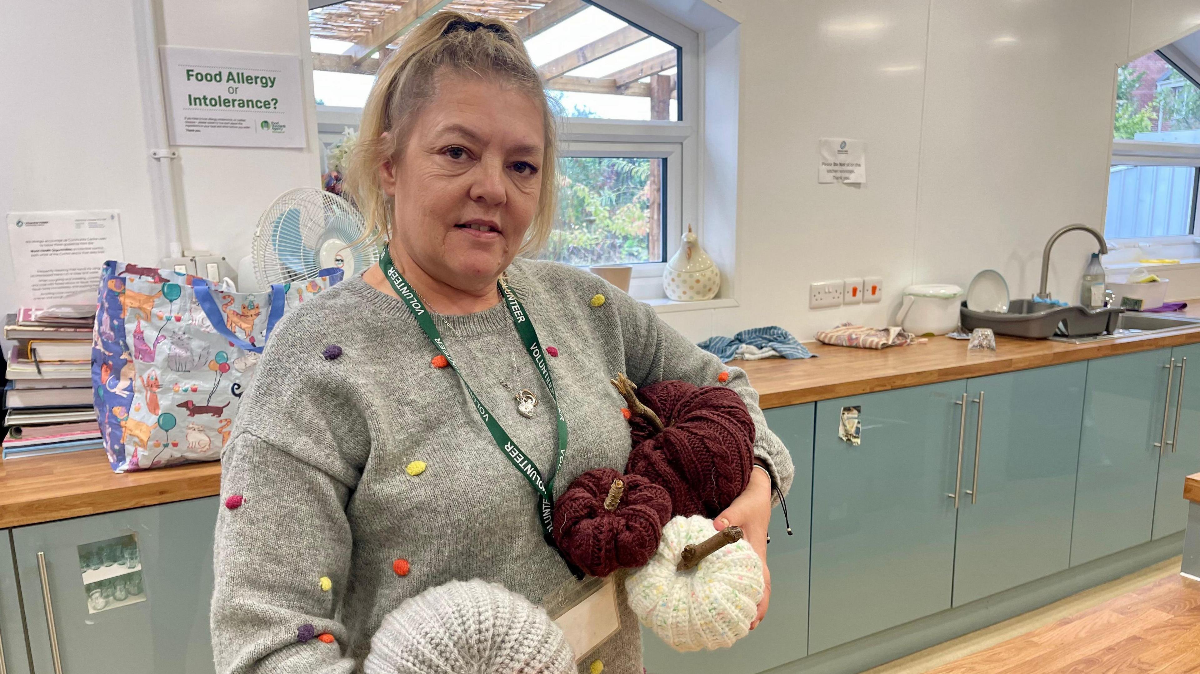 A woman in a grey jumper with small colourful dots stands in the kitchen of a community centre. She is holding knitted pumpkins of varying sizes and colours - maroon, white and grey. She has long, fair hair tied back in ponytail. She wears a green lanyard which saying "volunteer" in white. The kitchen has white walls, light wooden worktops, sage-coloured cupboard doors and light wooden flooring.