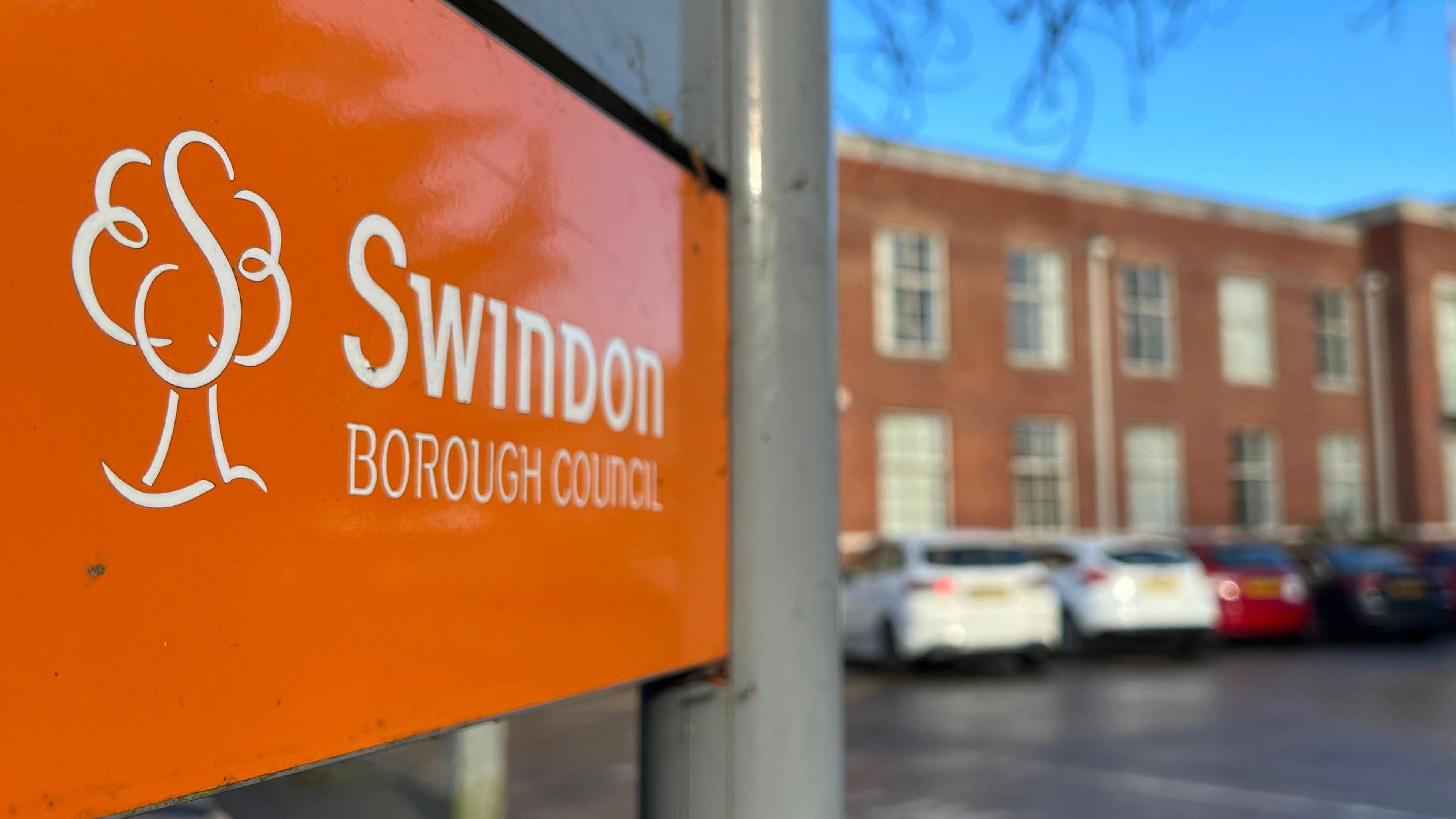 An orange Swindon Borough Council sign with a building behind it, with cars parked outside