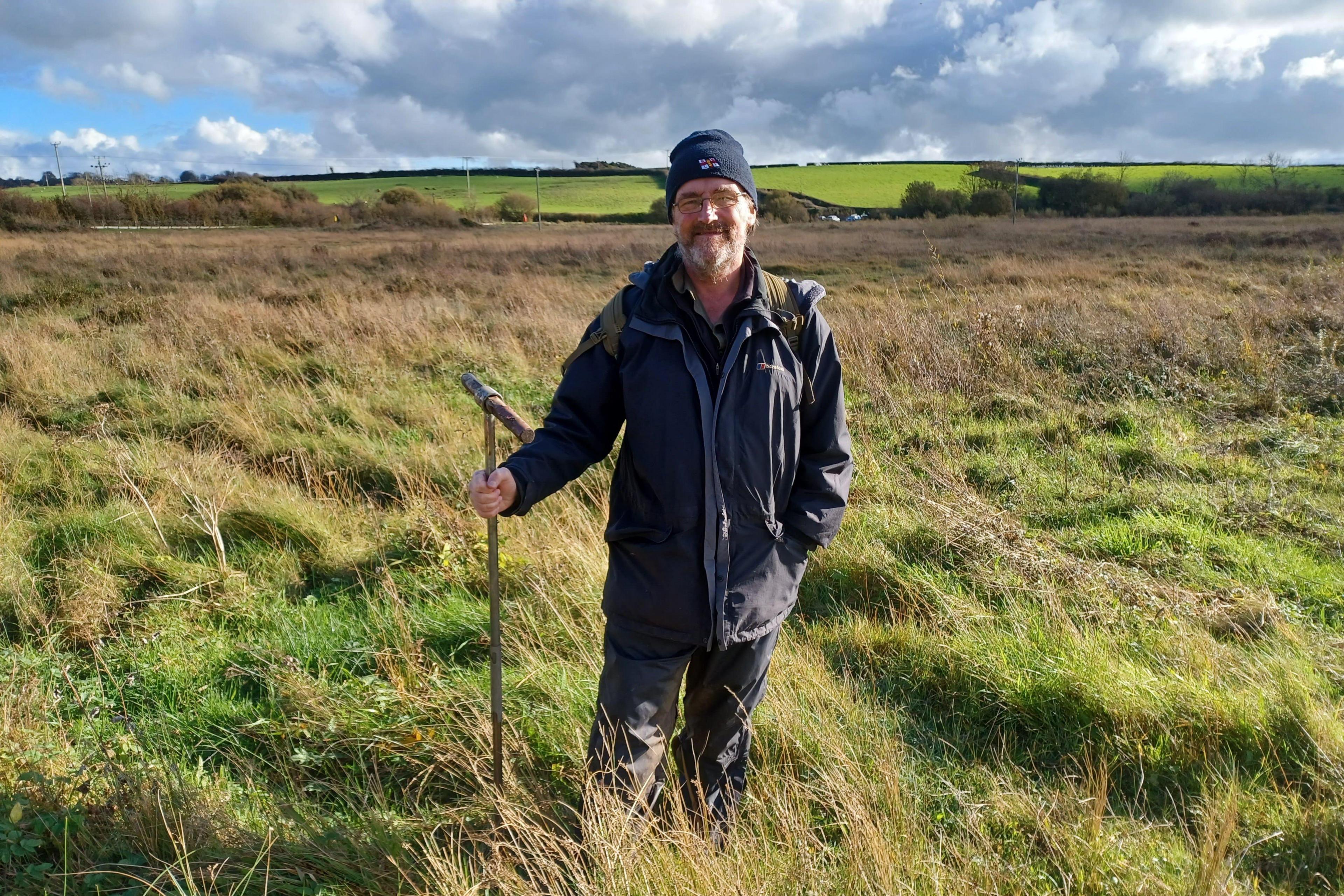 A person wearing outdoor clothing, a dark knitted hat and a backpack stands in a wide expanse of rough grassland, holding a long metal probe used for surveying peat or soil. Tall grasses surround the person, with shallow hollows and uneven ground visible in the foreground. Beyond the wetland, green agricultural fields and hedgerows stretch across gently rolling countryside. Large grey and white clouds dominate the sky, while bright sunlight breaks through to illuminate the vegetation, highlighting the contrast between the golden-brown grasses and the vivid green fields in the distance. The image illustrates fieldwork taking place in a peatland landscape.