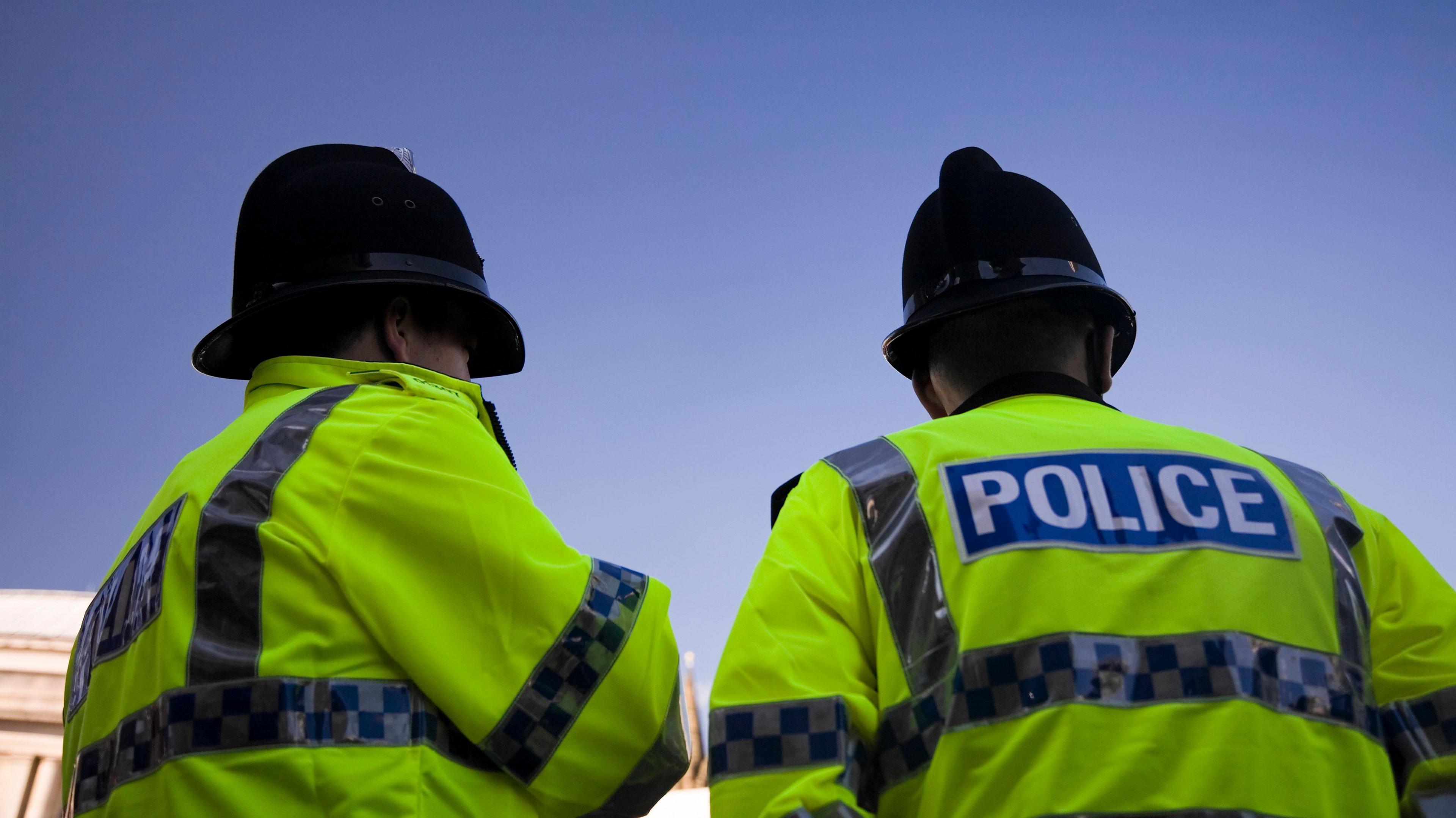 Image of two police officers. They are pictured facing away from the camera. They are wearing high vis police jackets and helmets