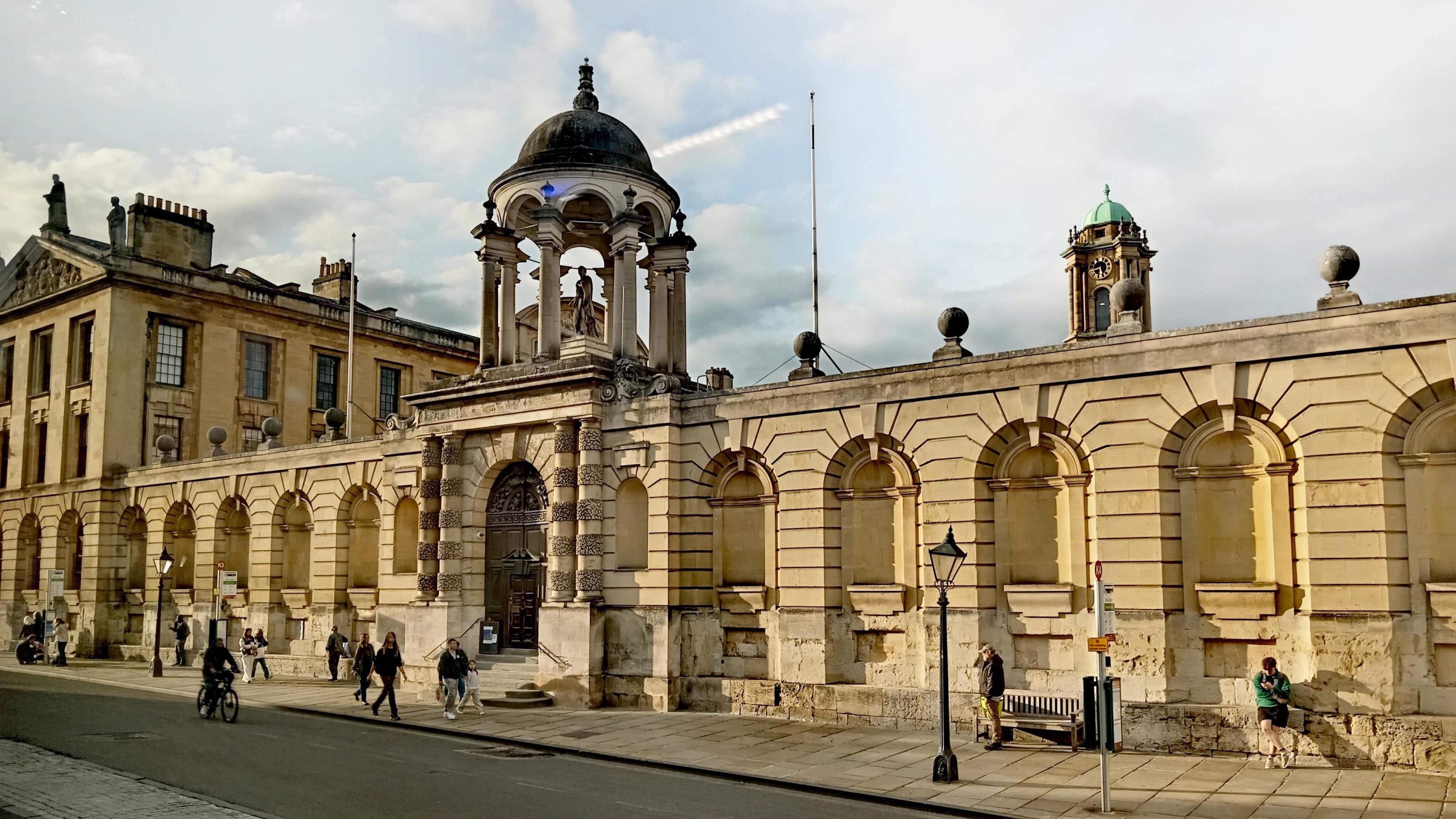 Old buildings with yellow coloured bricks. It has a tower with a dome on top and there are various statues visible on top of the buildings.