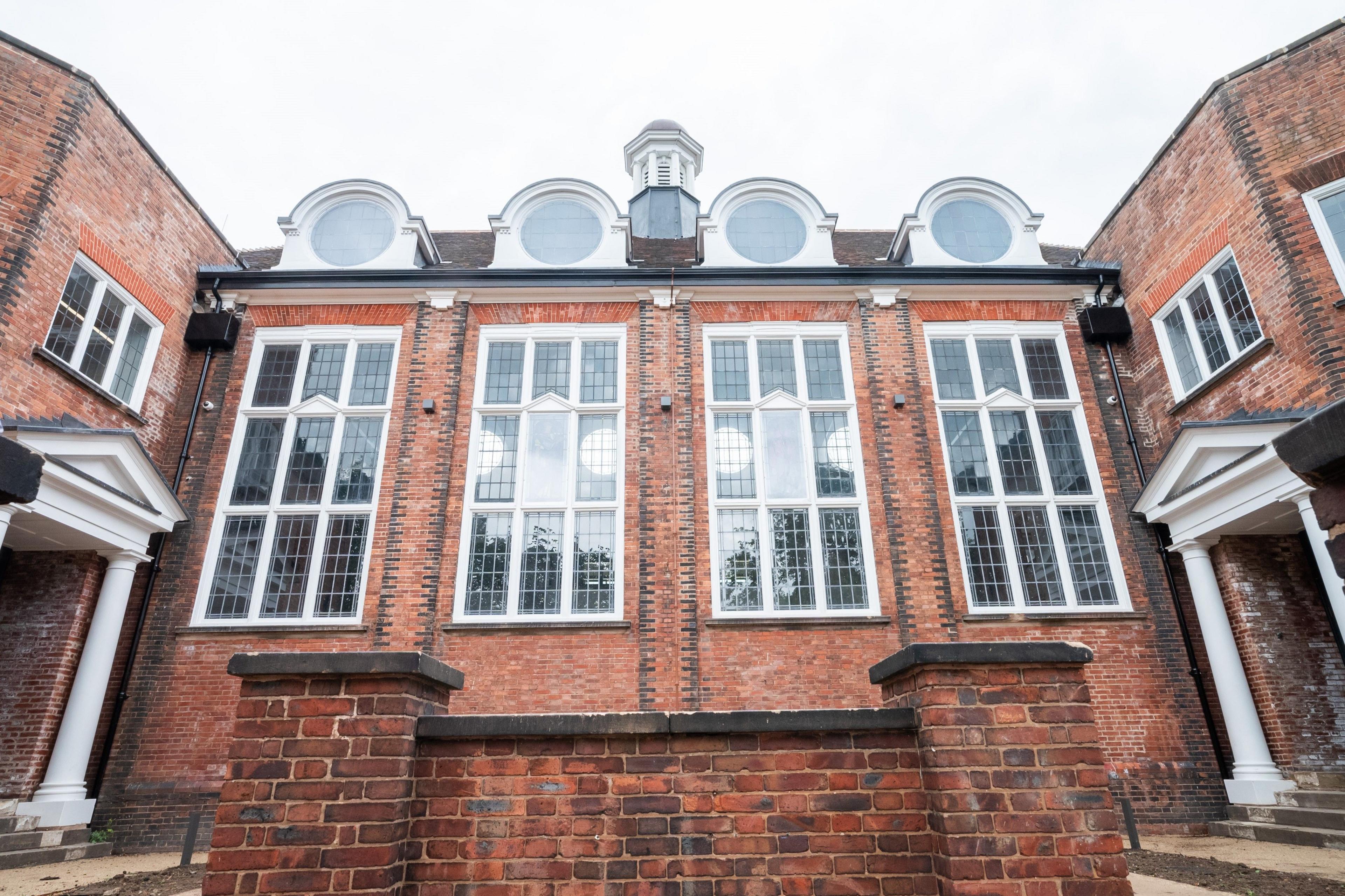 Close up of the front of the school. Red brickwork framing four large rectangular windows with a cupola and four circular windows above. Doorways with decorative porticos sit on either side.