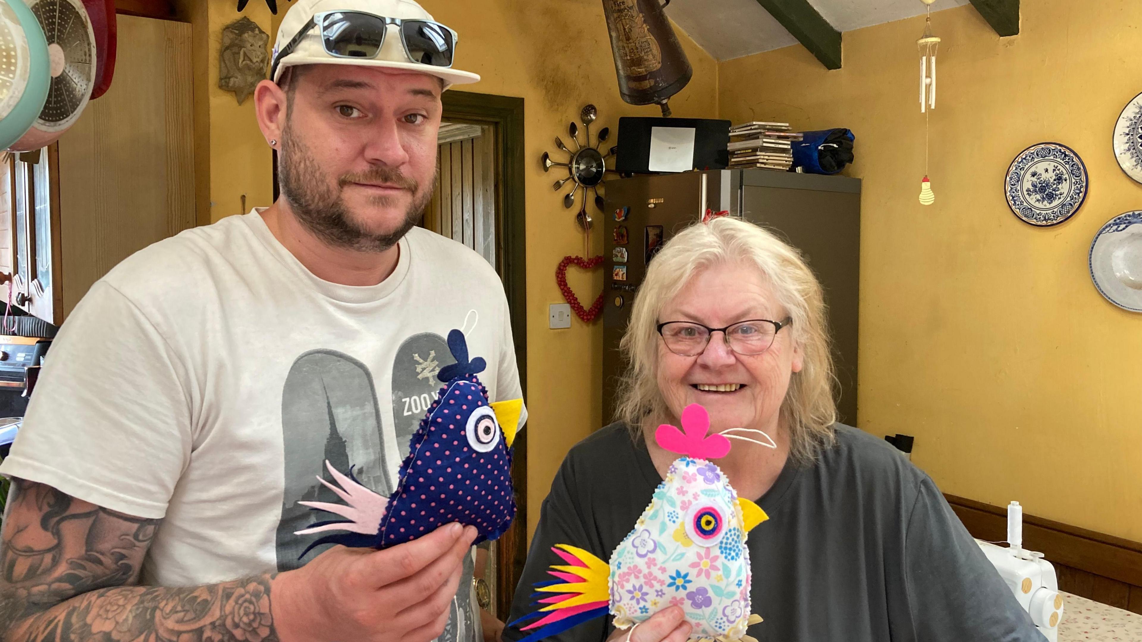 Sam on the left holding a purple and pink dotted, knitted hen and his mum next to him holding a multi-coloured chicken