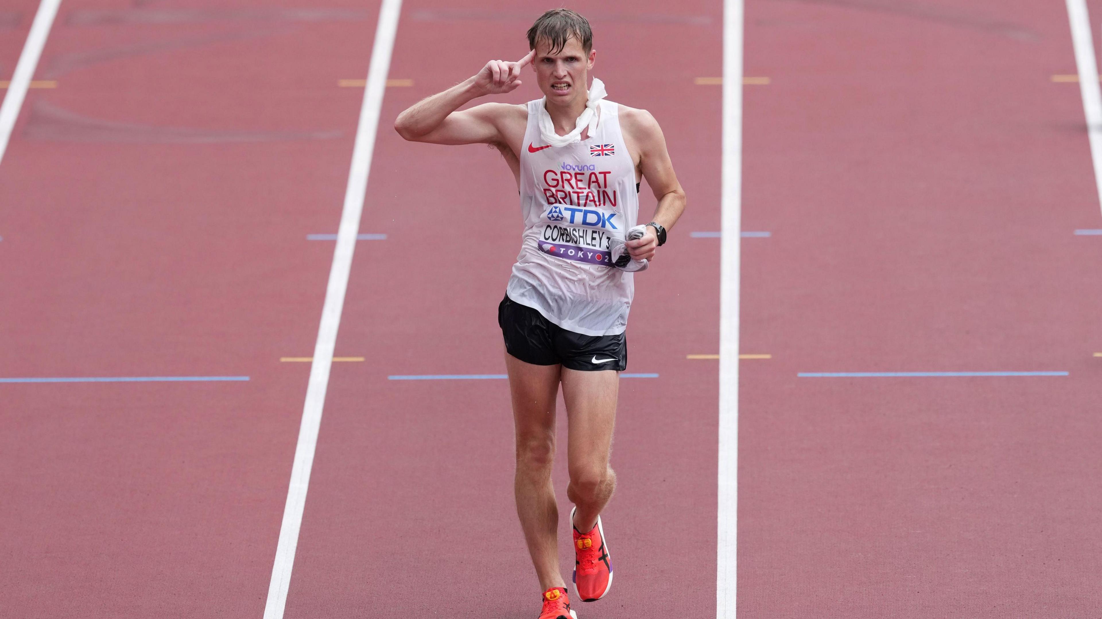An athlete is on an athletics track - he's wearing a white vest top, black shorts and orange trainers. He's wet, pointing at the side of his head, and looks exhausted
