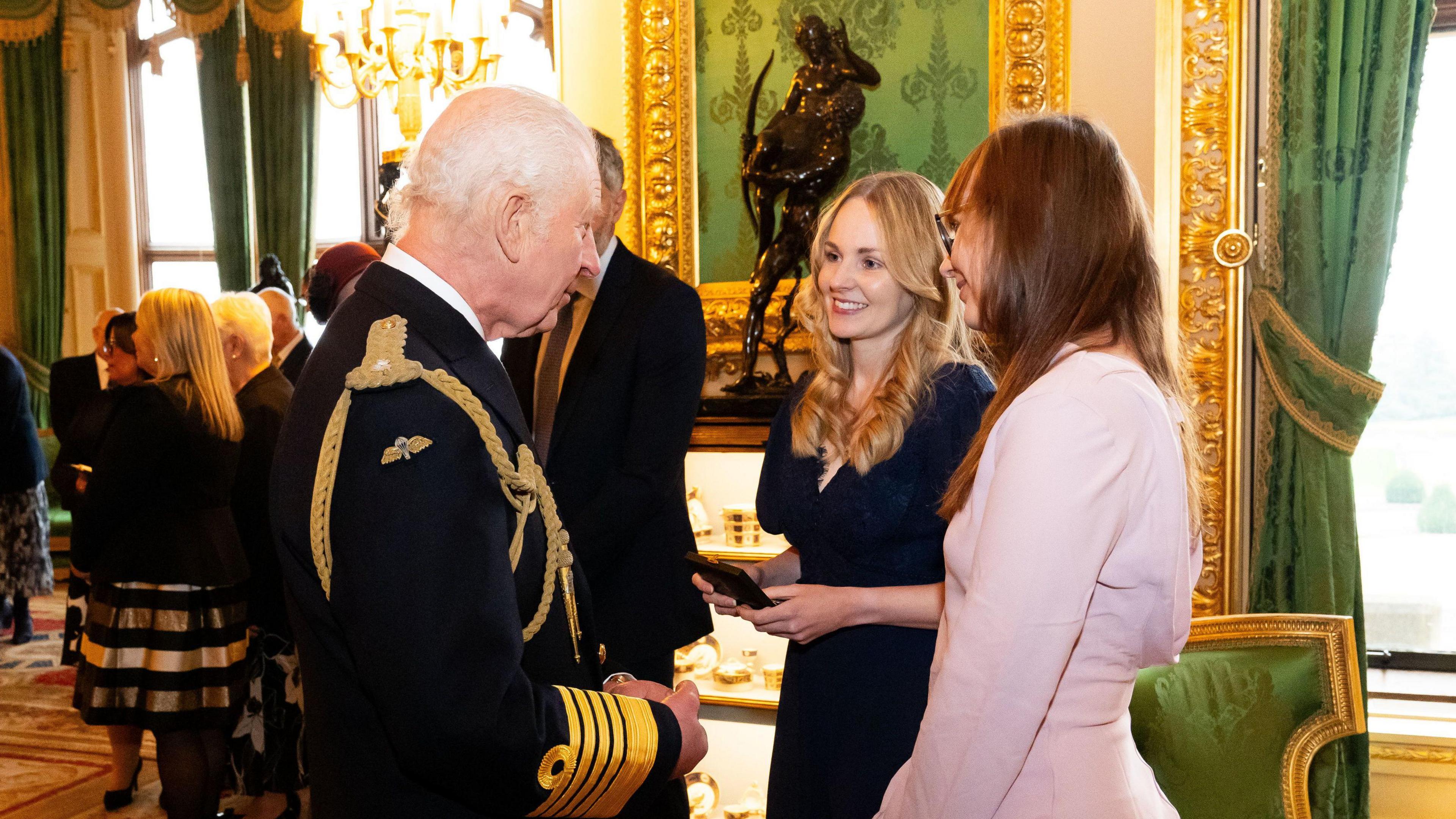 King Charles, in a navy blue gold lined ceremonial jacket, speaks to two women, one blonde and one brunette, in Windsor Castle