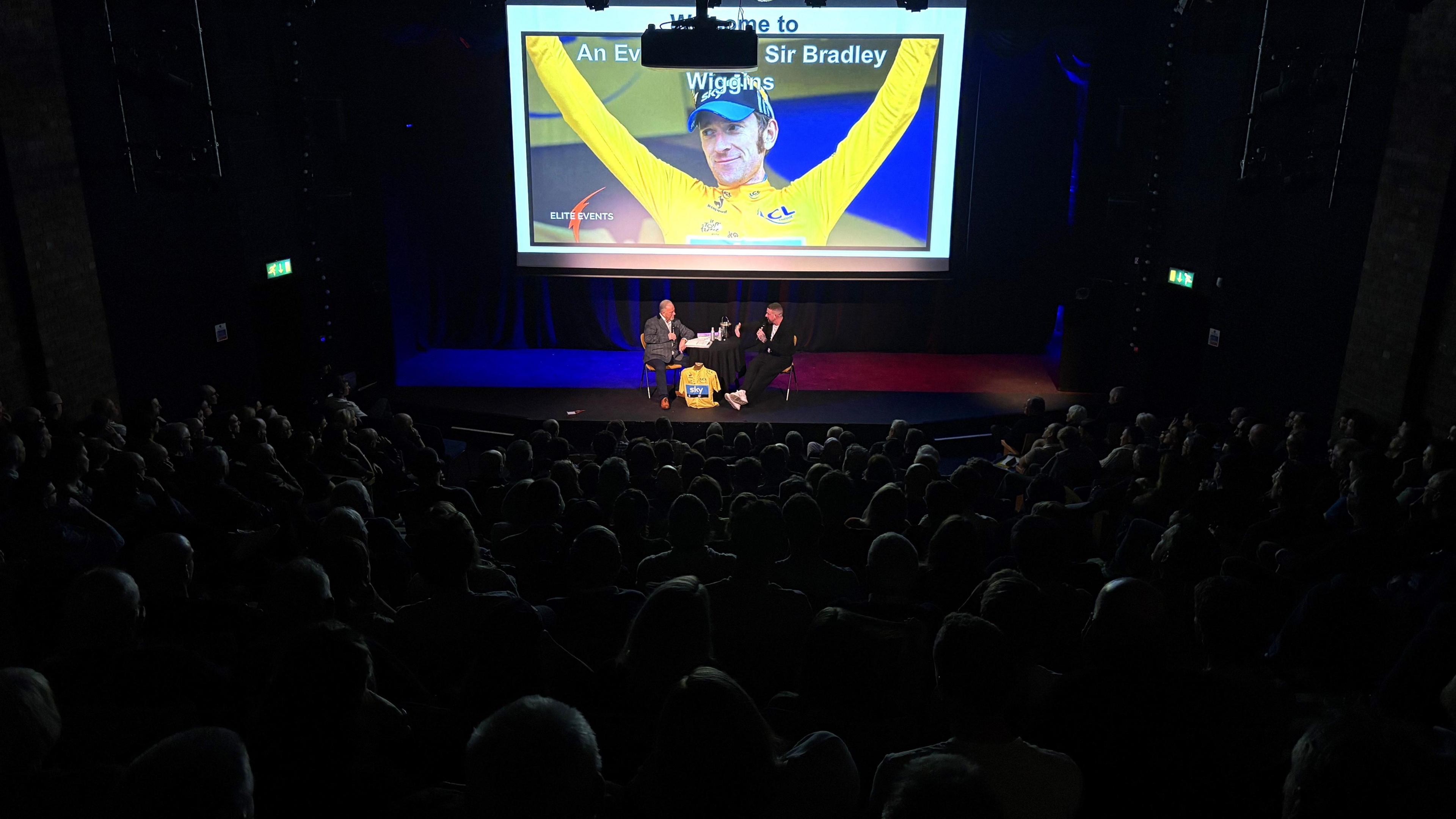 The main hall of the Redgrave Theatre in Bristol is viewed from the back, with the seats filled with audience members but in darkness. On the stage are two men facing each other sitting down, and above them is a large screen with a picture of Sir Bradley Wiggins wearing the yellow jersey of a Tour de France winner