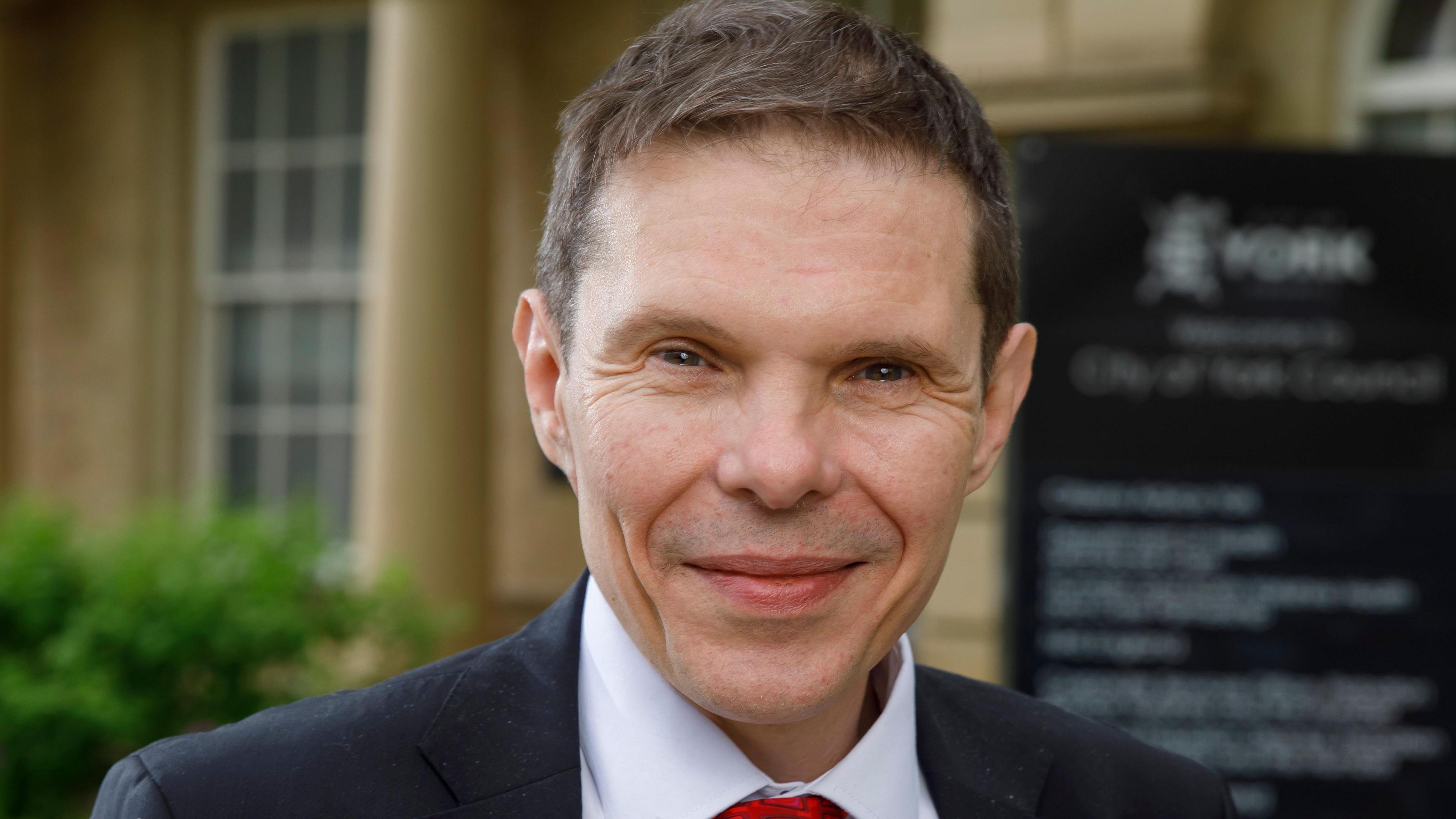 Councillor Pavlovic in the centre of the screen, smiling. He has brown hair and is wearing a suit. 