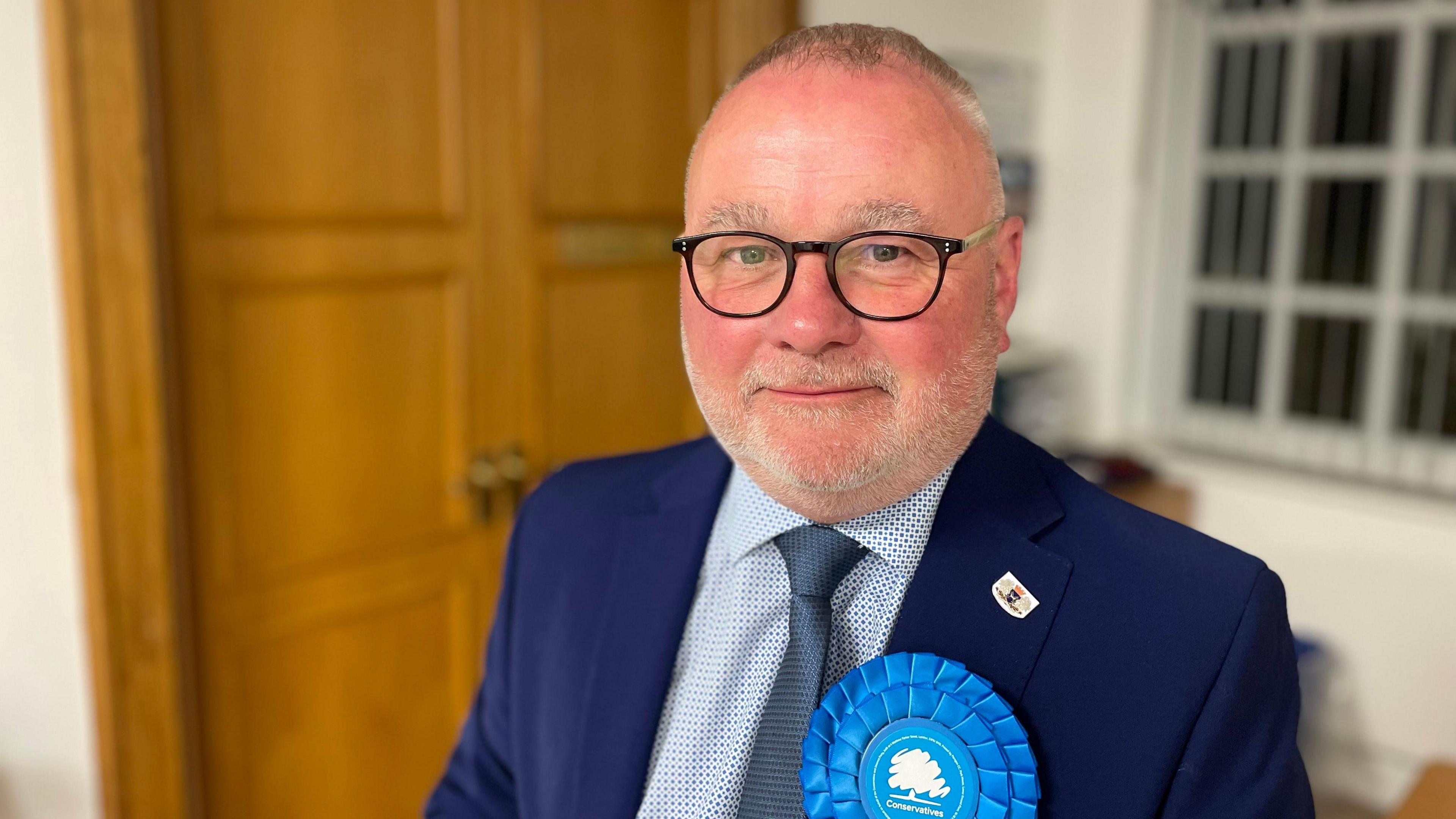 Conservative group leader Wayne Fitzgerald wears a suit and blue Conservatives rosette. He is standing in front of wooden doors. 