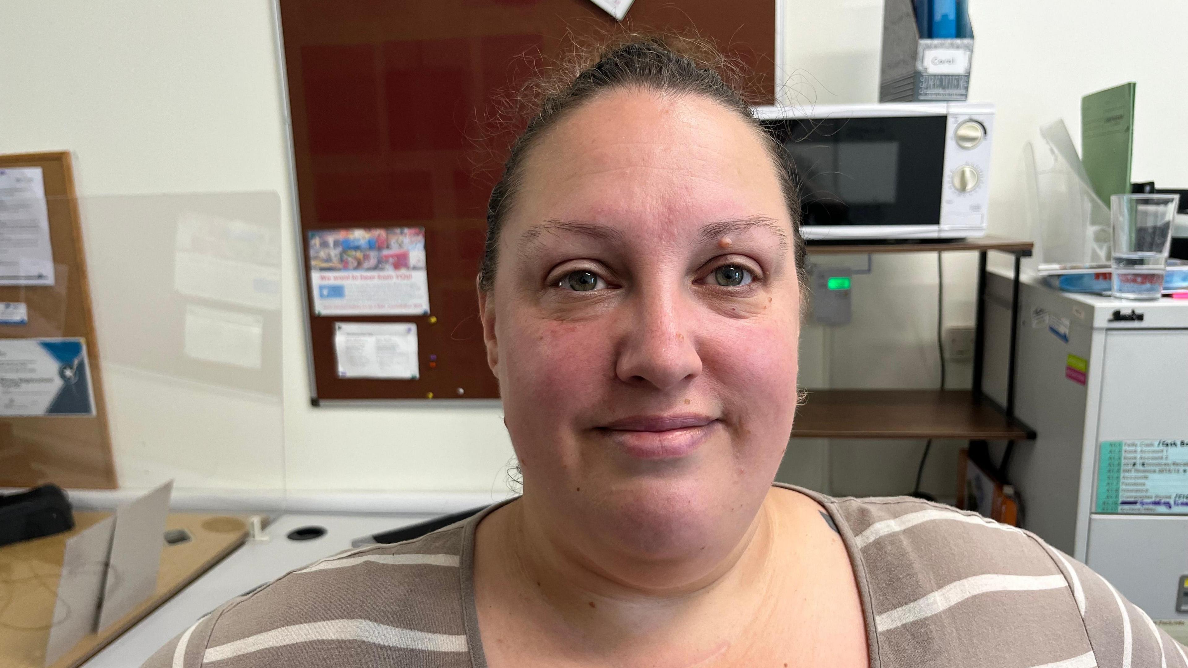 Jodie Montgomery, 40, has black high pulled tightly over her head, she ahs a light brown and crème striped jersey on and behind her are items in an office, a noticeboard and a microwave and part of a tall filling cabinet.