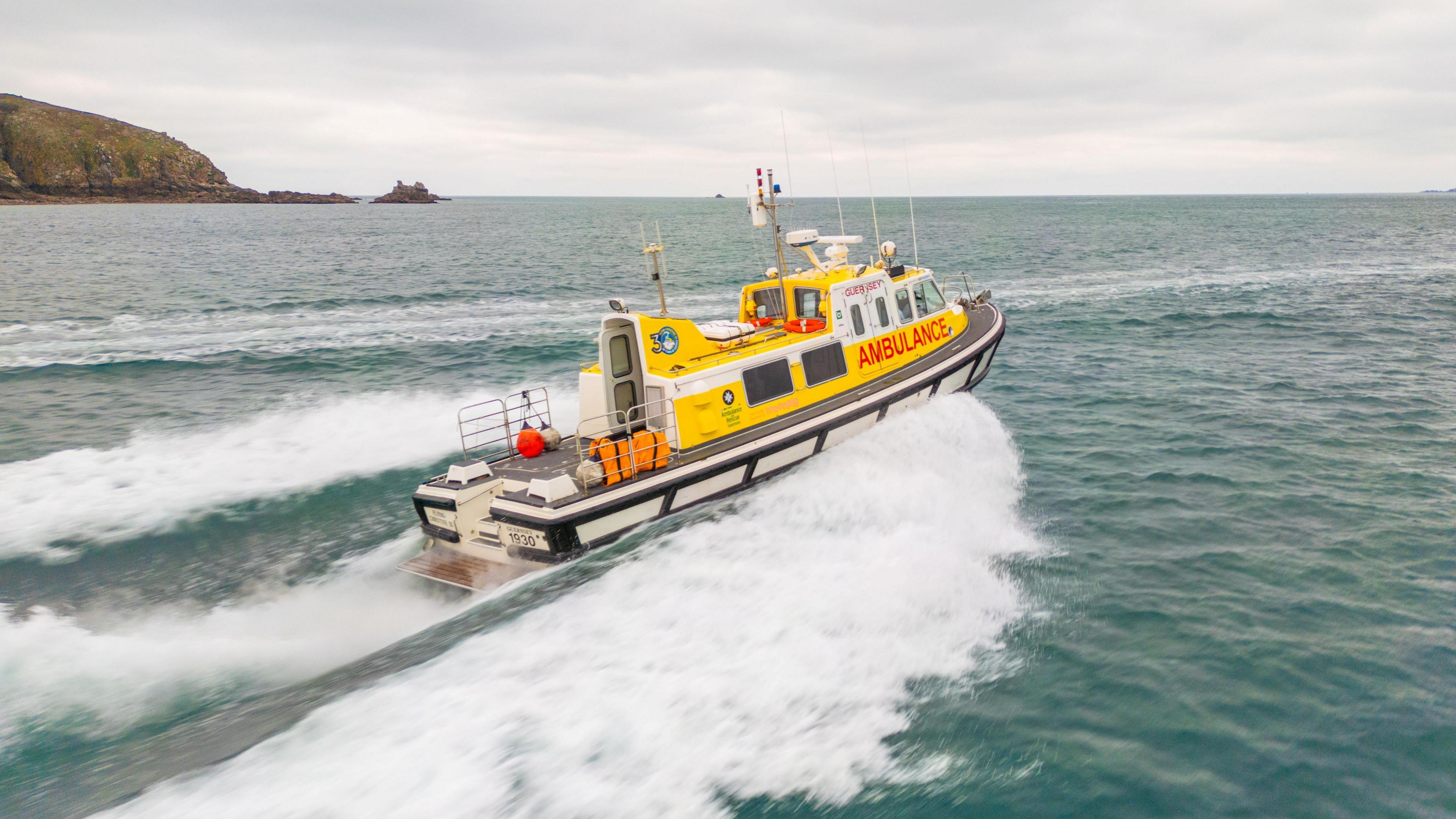 Marine ambulance speeding through the sea. The boat is yellow, white and black. Guernsey and ambulance are written in red and its name Flying Christine III is written in white.