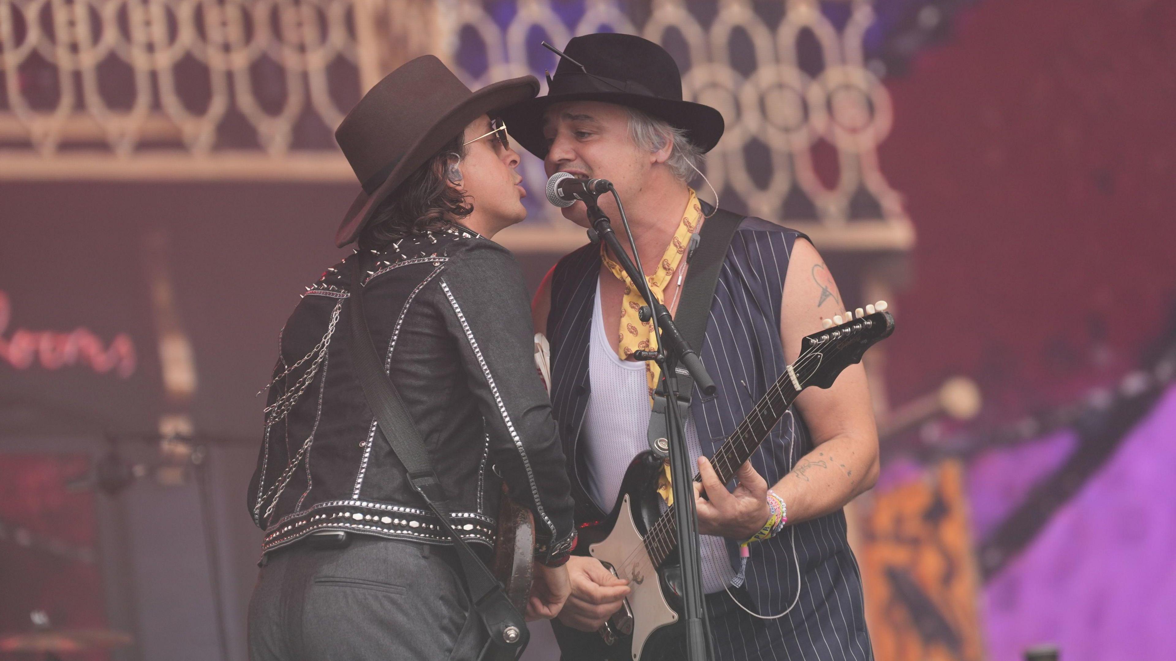 Two people are sharing a microphone while playing the guitar. They are both wearing hats.