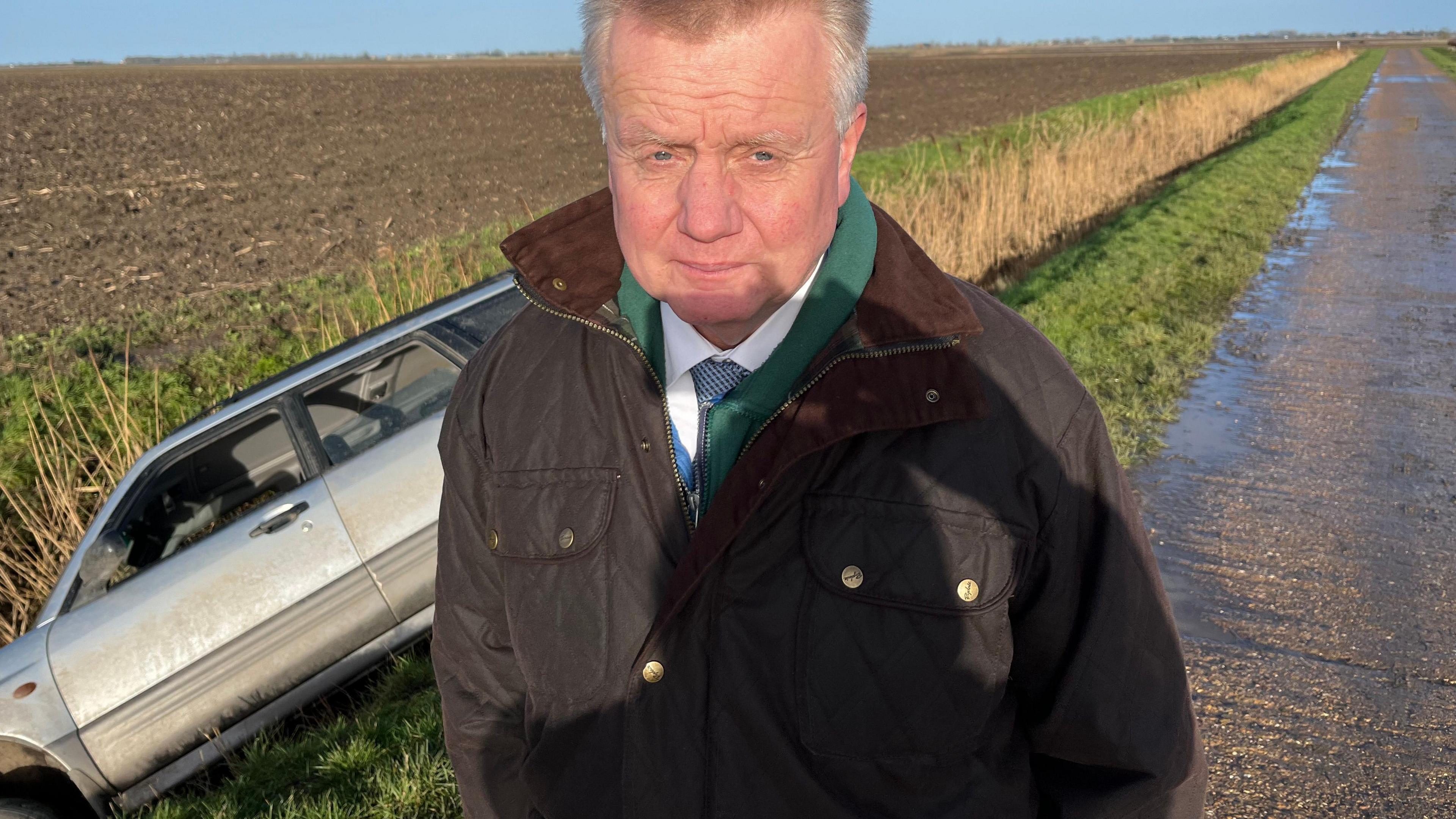 Man stands on side of a road next to a ditch. A car be seen lying in the ditch. Fields surround the road and ditch.