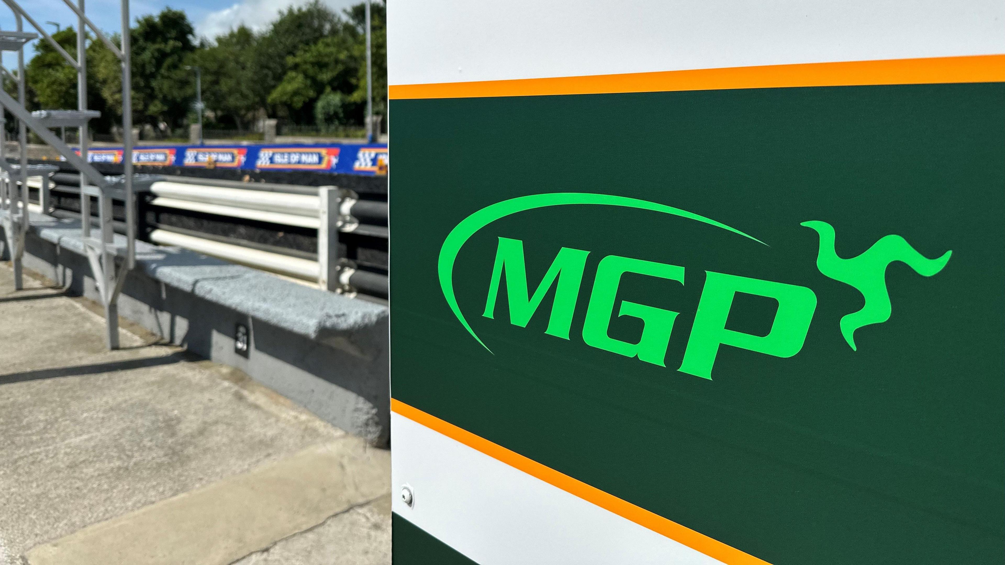 A dark green sign with the MGP logo on it in the pit lane. A blue and red banner is hanging from the barrier across the road in the background. 