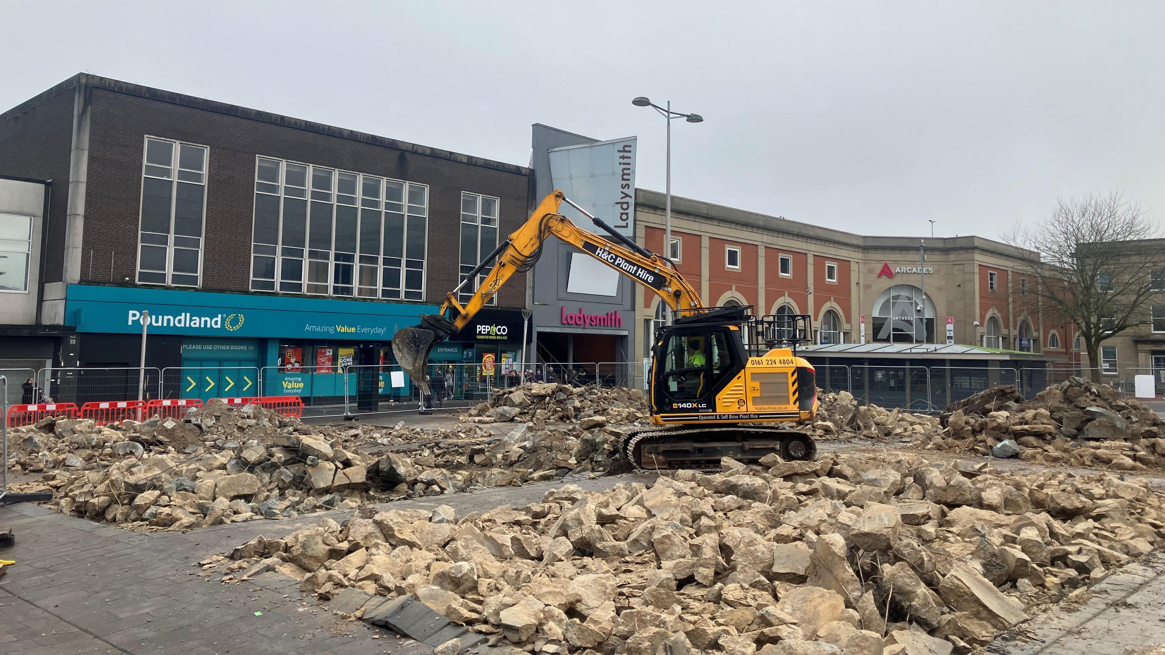 Photograph of a bulldozer in Ashton-under-Lyne. Large piles of broken concrete lie in them middle of an open space in front of Poundland.