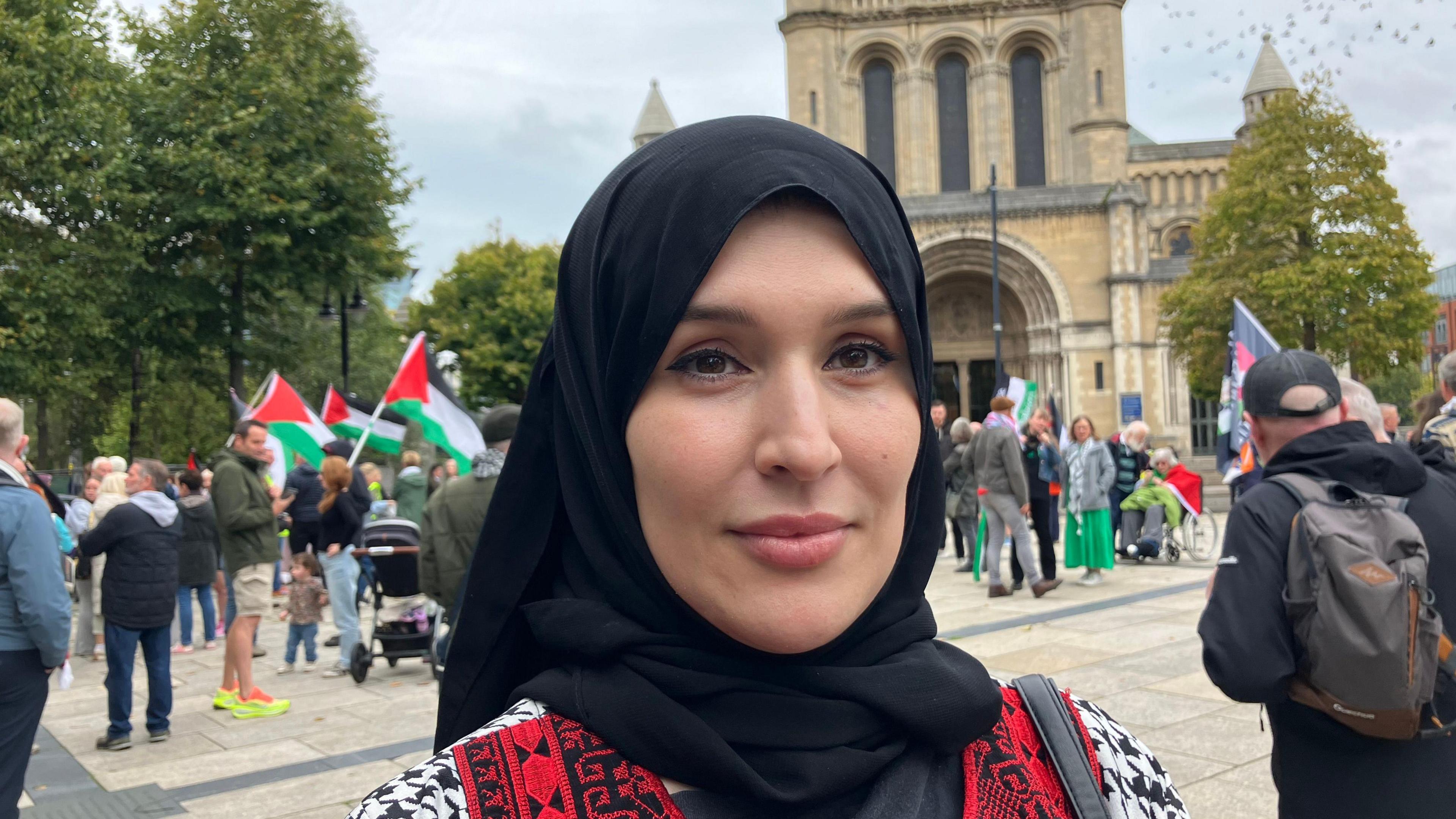 Eftaima Najjair at the protest in Belfast, with St Anne's Cathedral in the background. She is wearing a black hijab scarf on her her and a black and white patterned coat with red and black panels at the front. Protesters holding Palestinian flags are standing in the distance behind her.