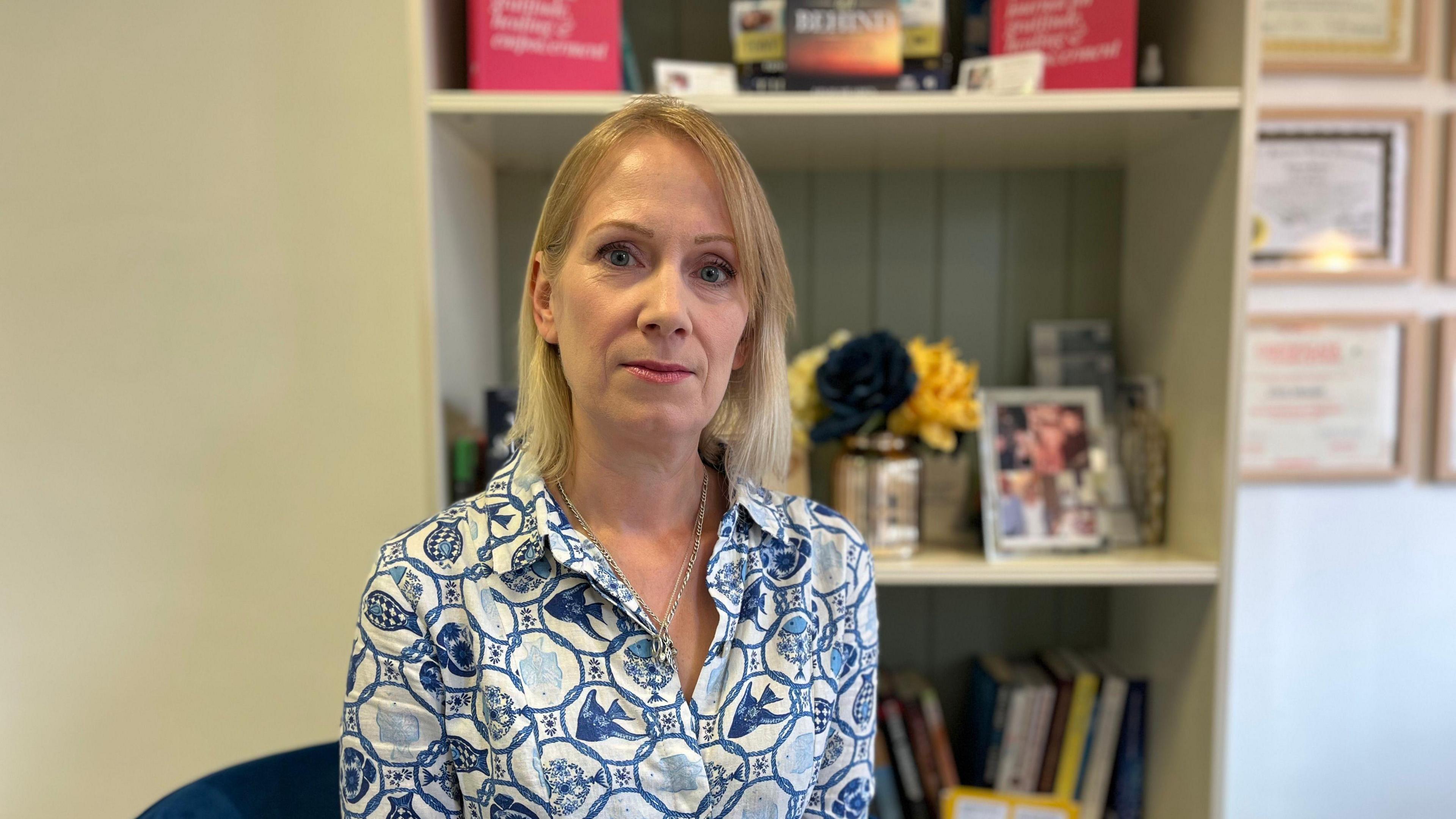 A woman wearing a shirt with a blue and white pattern sat in her home, in front of a bookcase. She has a stern expression.