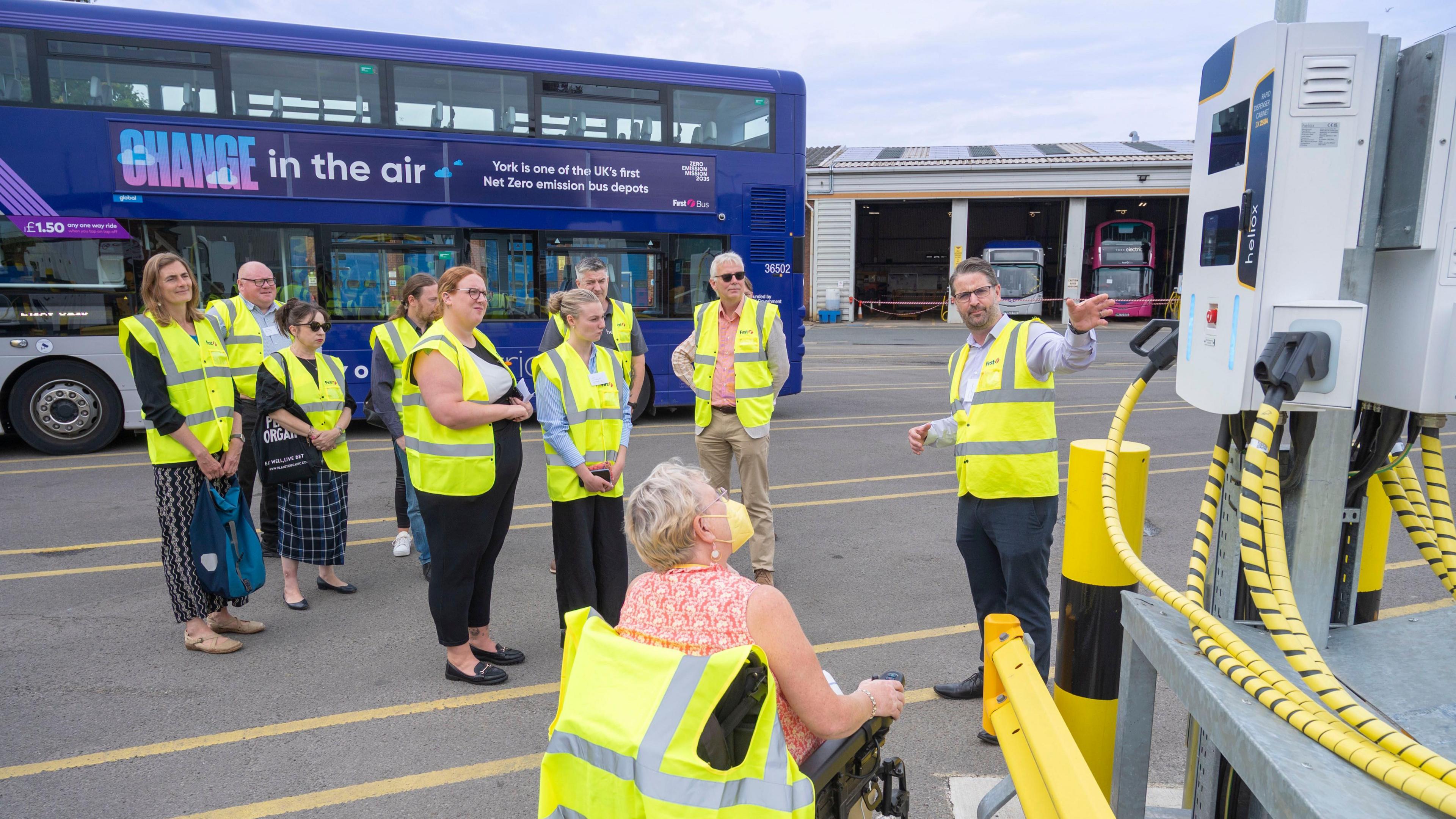 A group stands in hi-vis jackets in a York bus depot. They are stood in front of a blue First bus as a man shows them an electric charging point