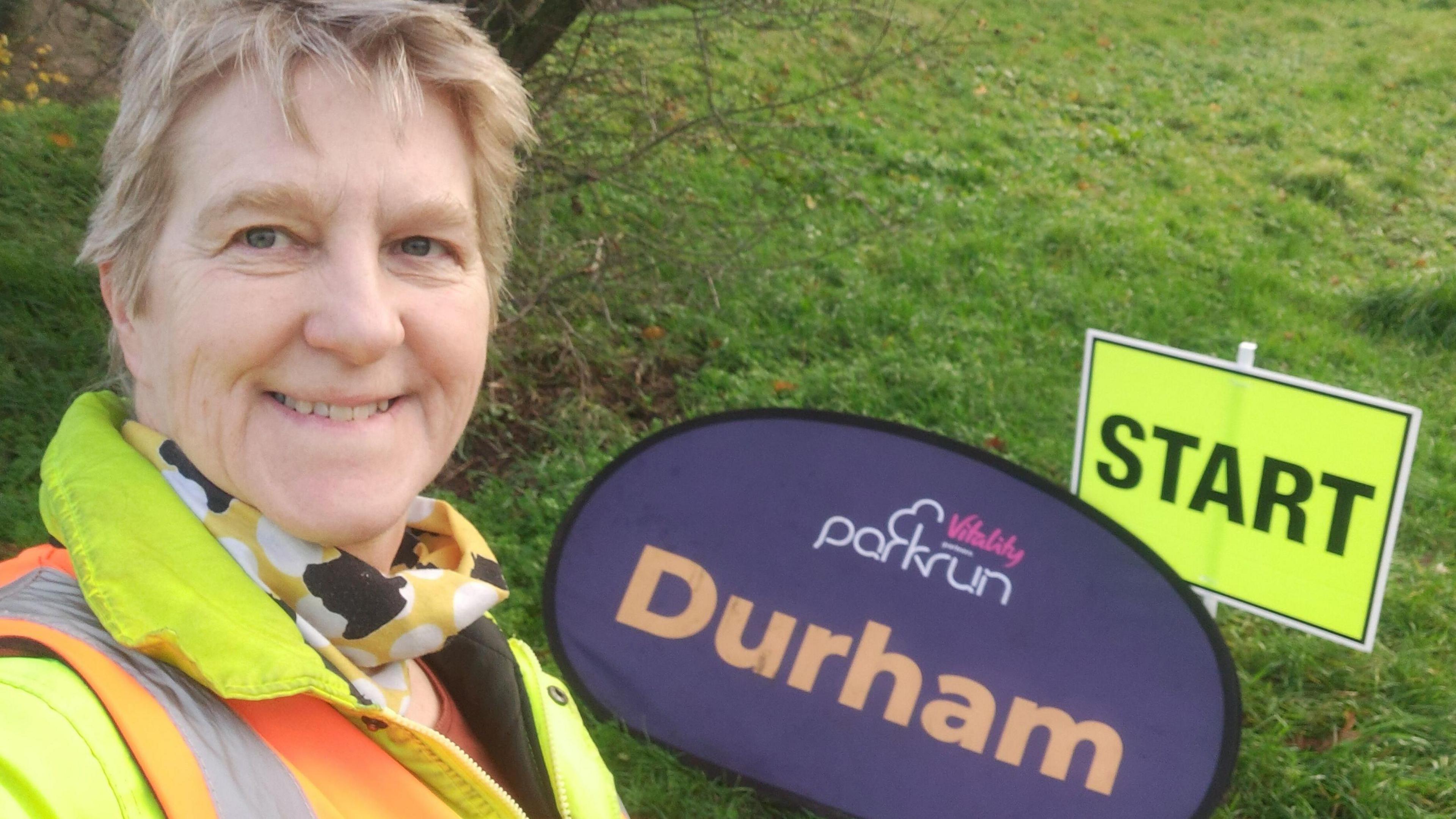 Amanda poses with Durham sign at parkrun event. She is wearing a fluorescent jacket.