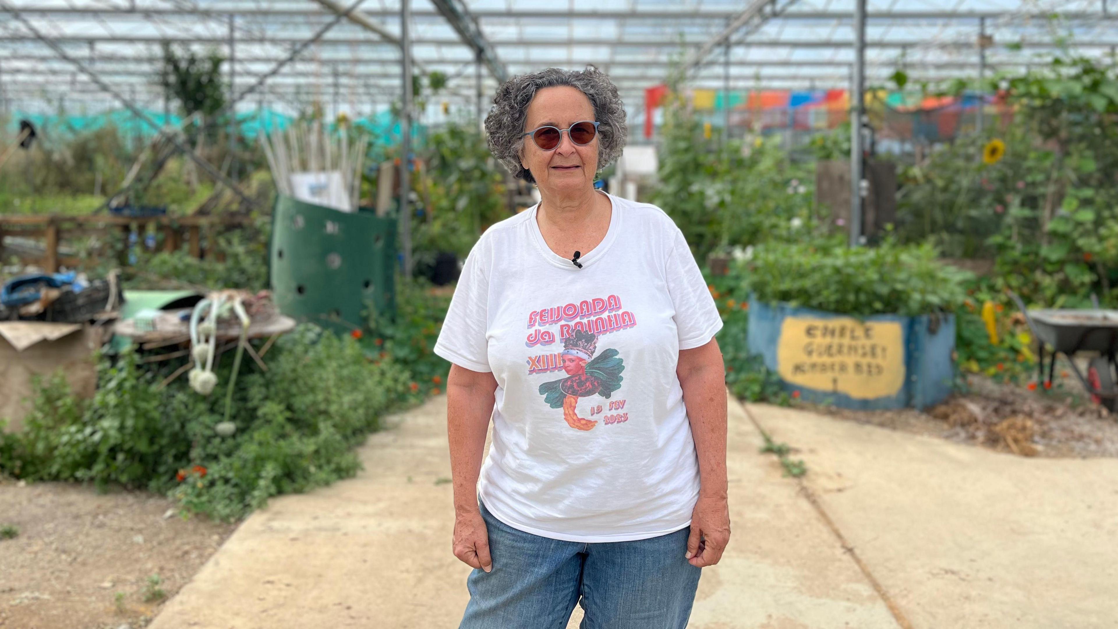 A photo of a woman standing in a greenhouse. She has short, curly, grey hair and is wearing sunglasses. She is wearing a white t-shirt with red writing on and blue denim jeans.