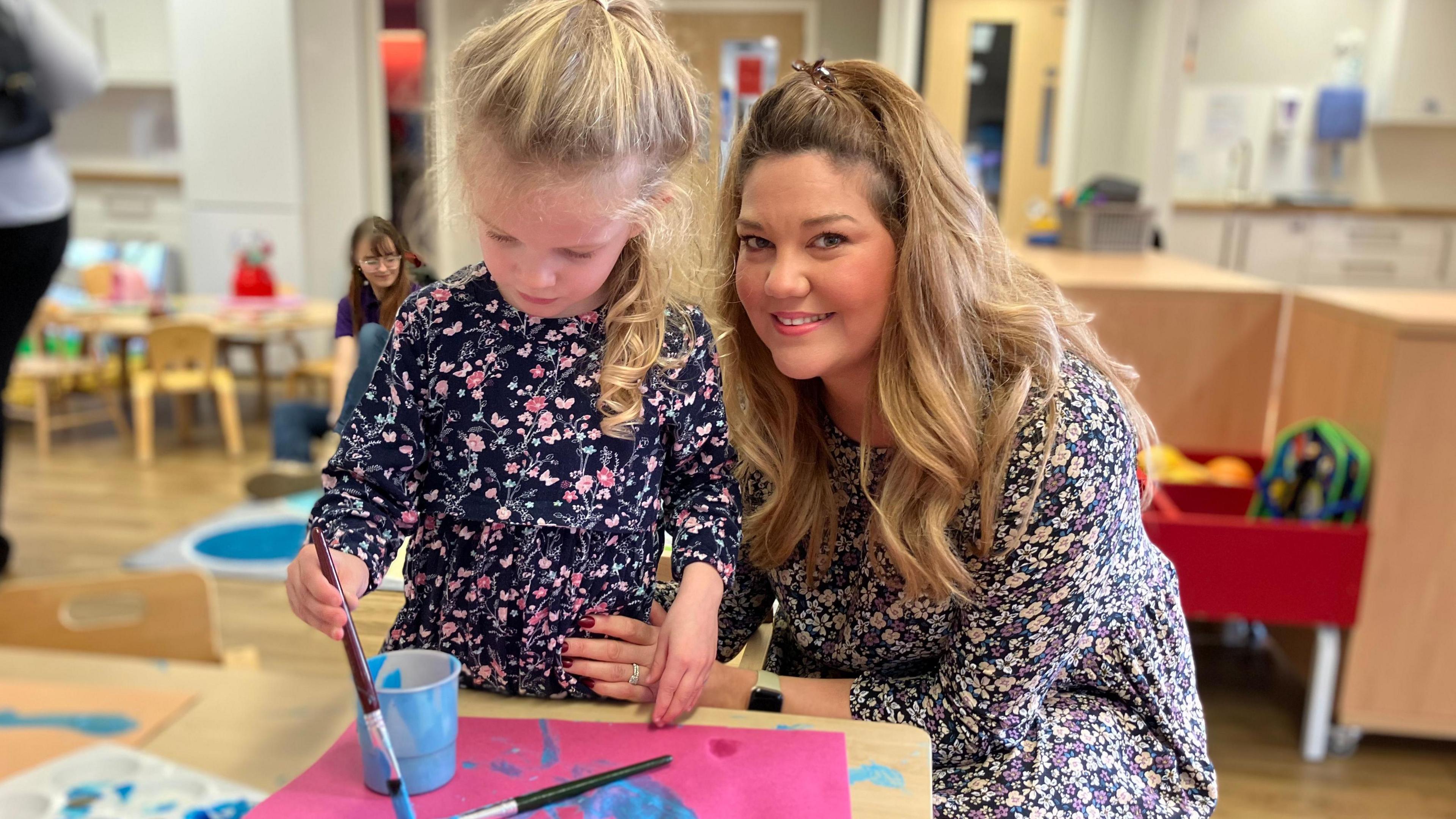 A girl with blonde hair painting while her mum, also with blonde hair, cuddles her.