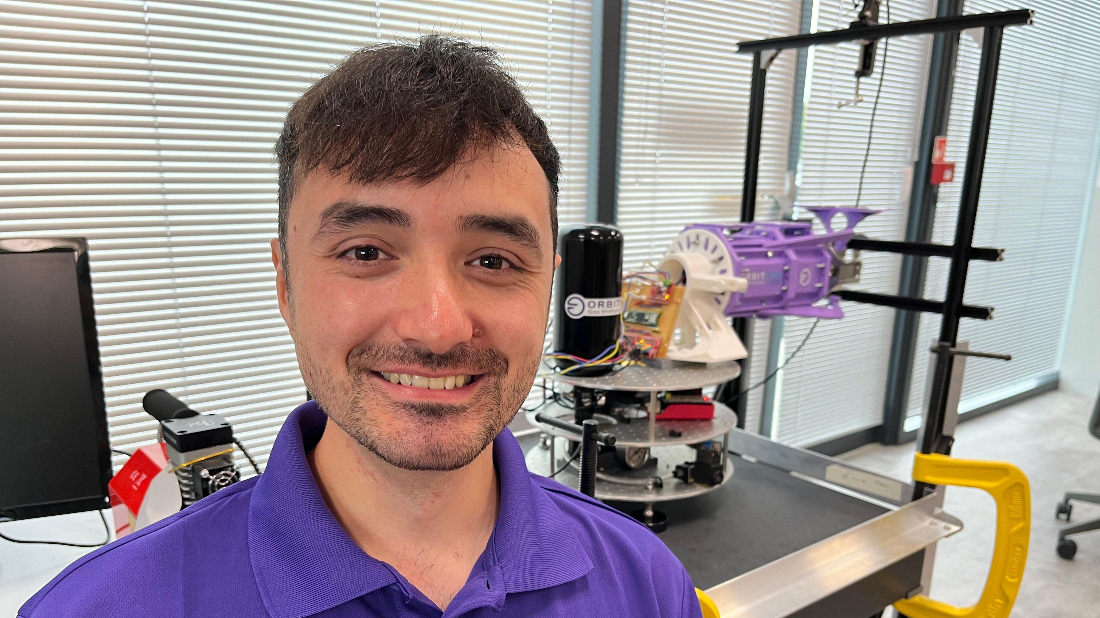 Omar Raj is wearing a purple polo shirt and has short brown hair and brown stubble. Behind him is part of the technology that Orbit Fab are developing.