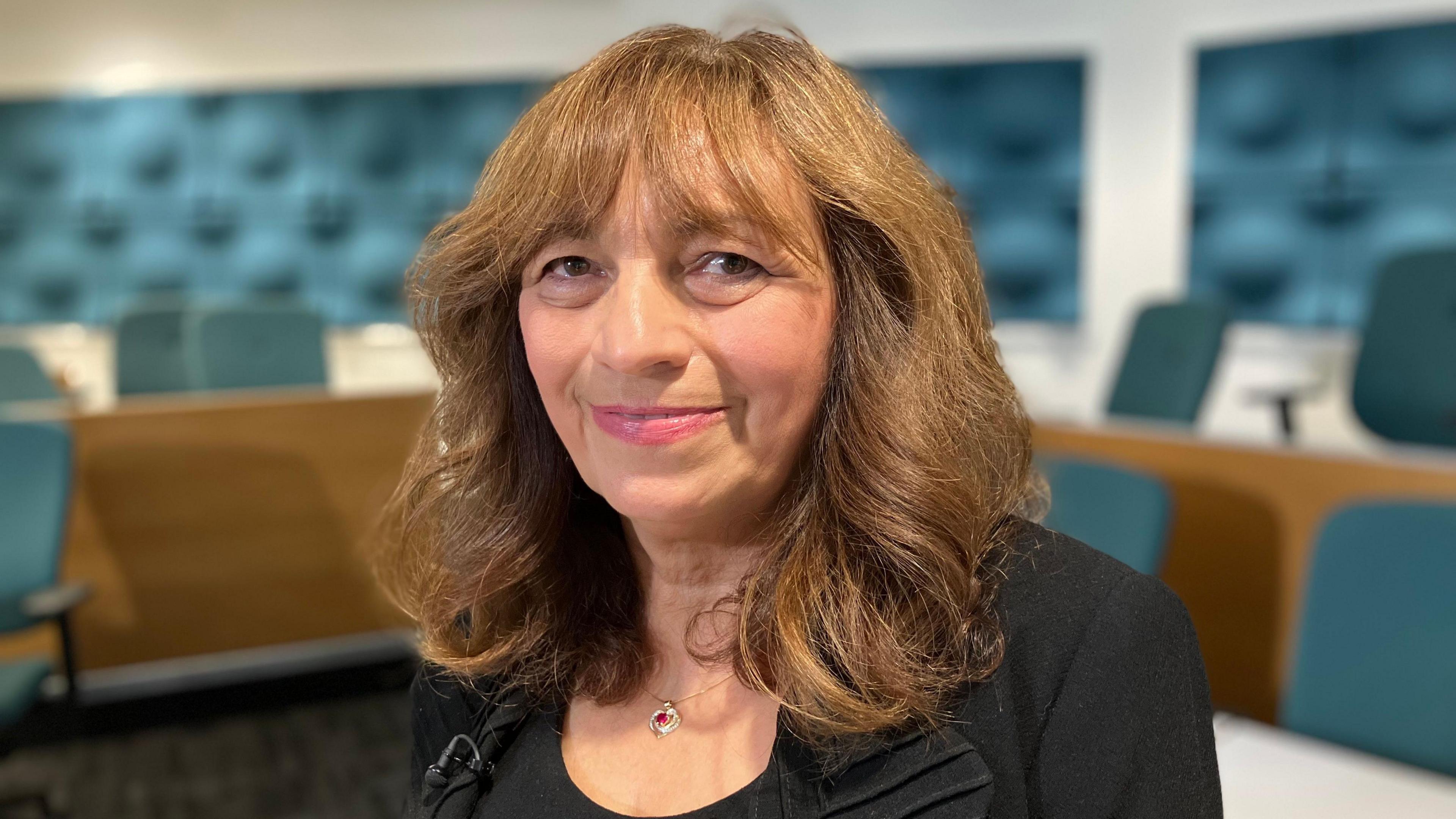 A head and shoulders picture of Christine Quinlan who is looking at the camera and smiling. The background is blurred but she is standing in front of wooden desks that have green office chairs behind them. There is green textured panelling on the wall.