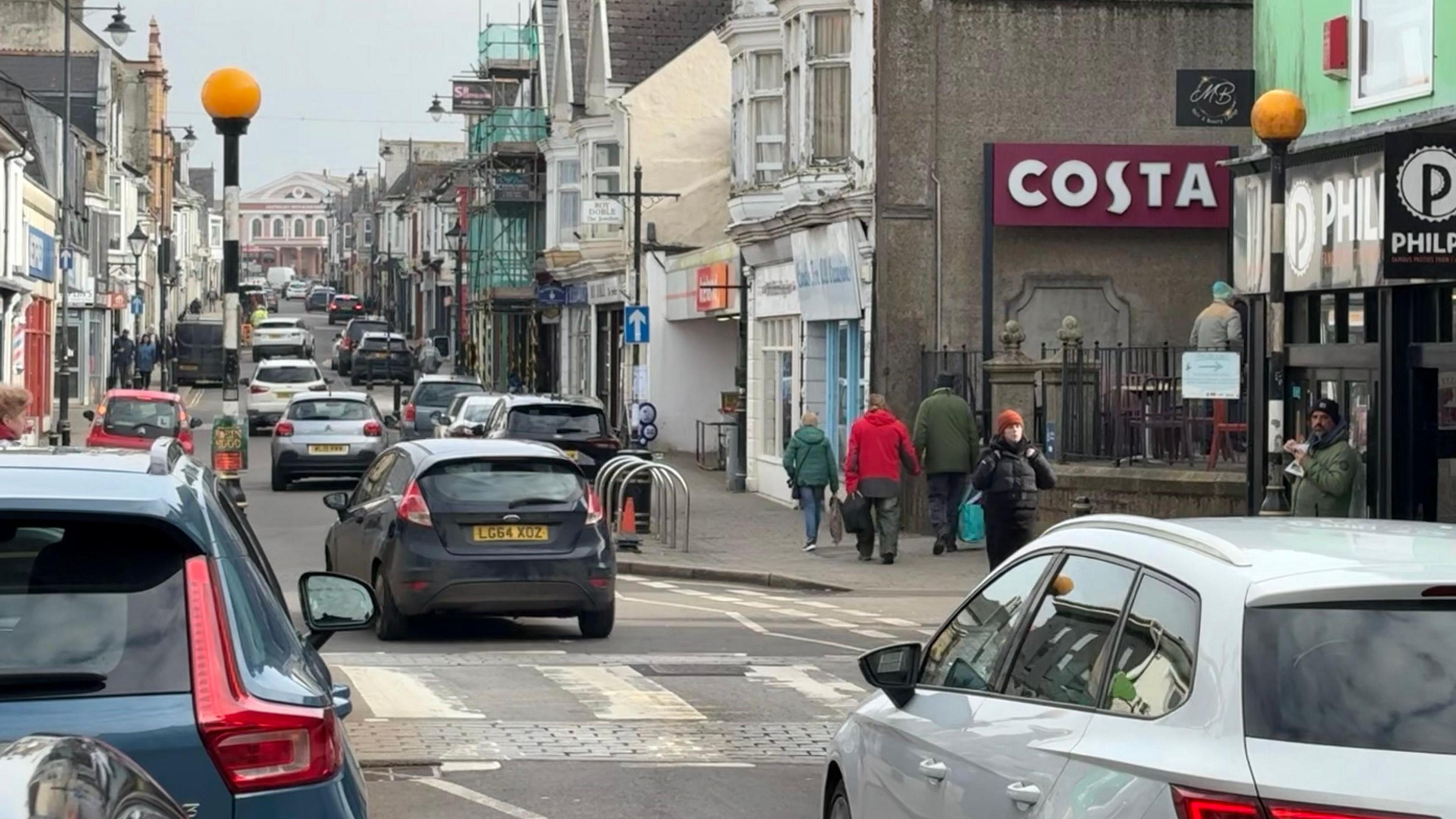 Trelowarren Street in Camborne. There are cars parked on either side of the road outside shops while other vehicles drive up the road. It is a busy scene with a number of people walking on the pavements.